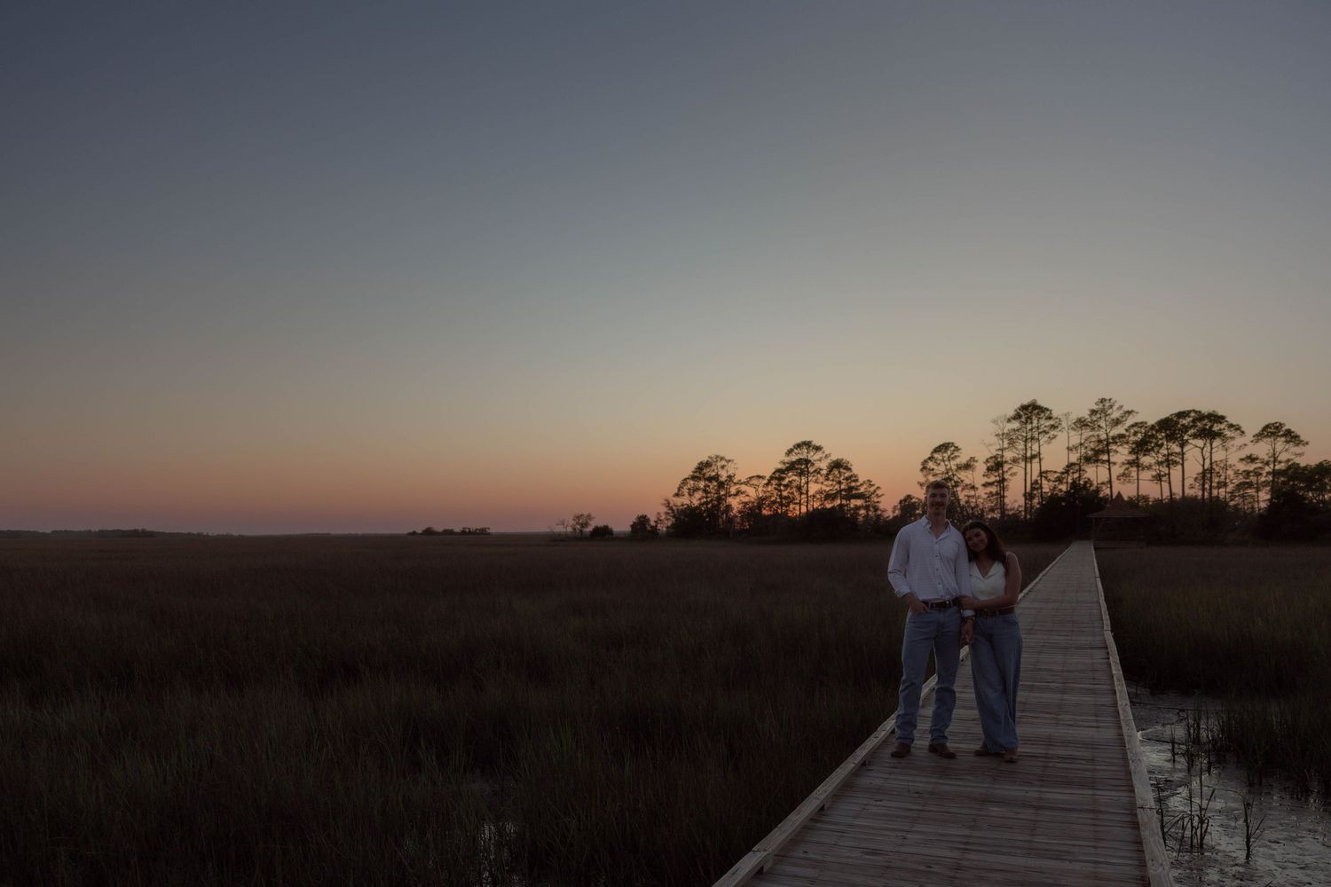 Couple standing on wooden boardwalk overlooking marsh at colorful dusk sunset with silhouetted pines.