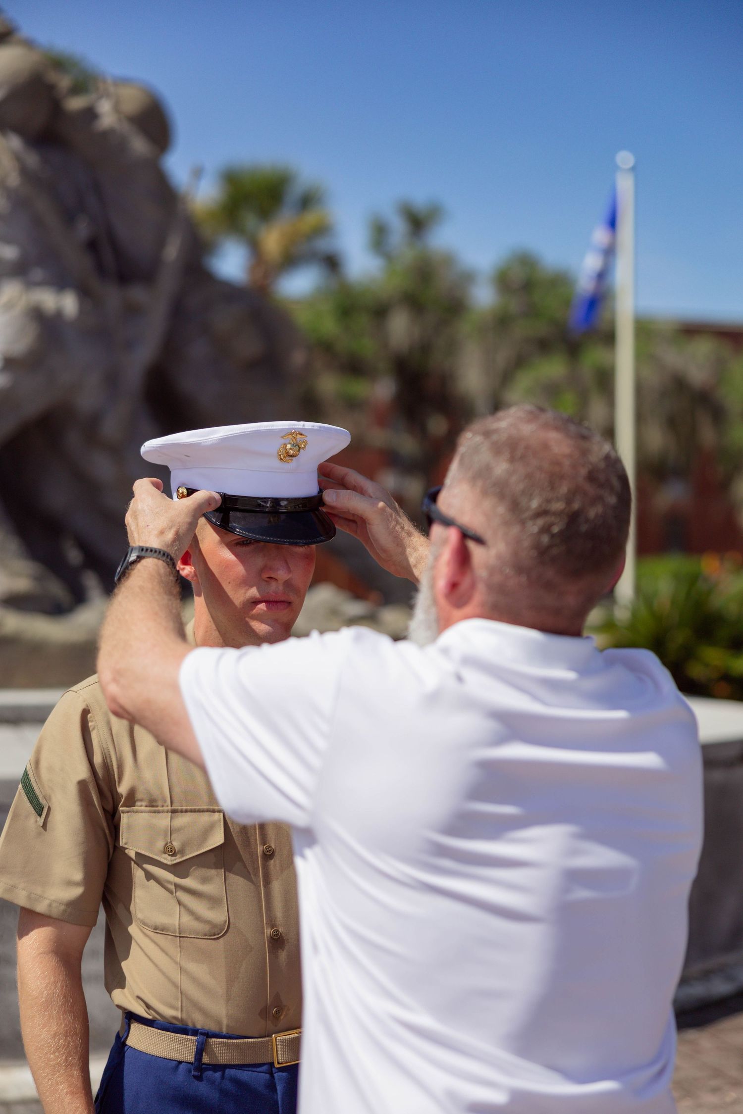 An older man in a white shirt carefully places a white dress cap on a young Marine in uniform outdoors.