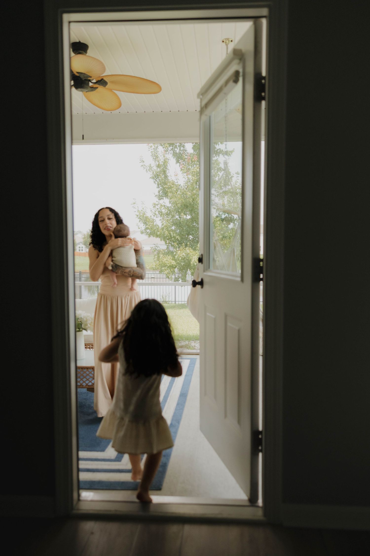 A young girl runs toward her mother holding a baby on a bright porch, viewed through an open doorway.