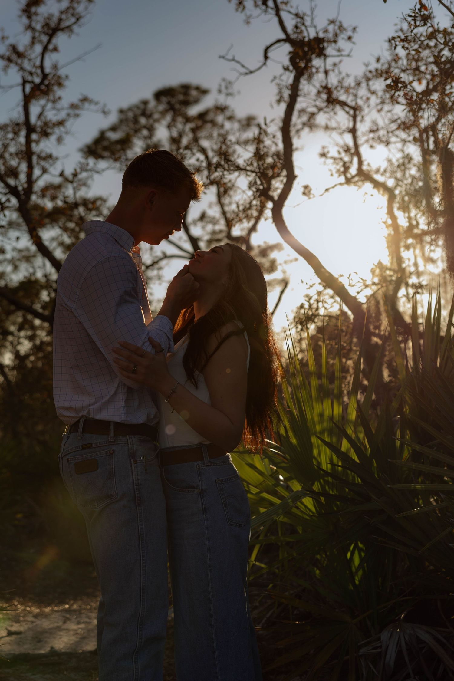 Romantic couple embracing in golden backlit sunset glow among tropical trees and lush greenery.