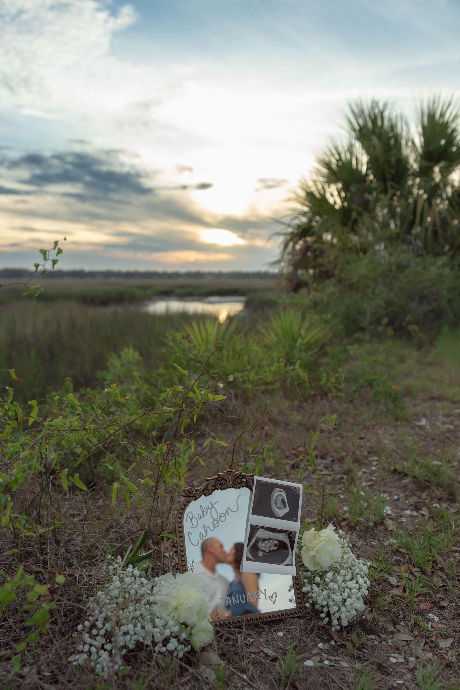 Sunset memorial display with ultrasound photos, baby name sign, and white flowers in a marshy landscape.