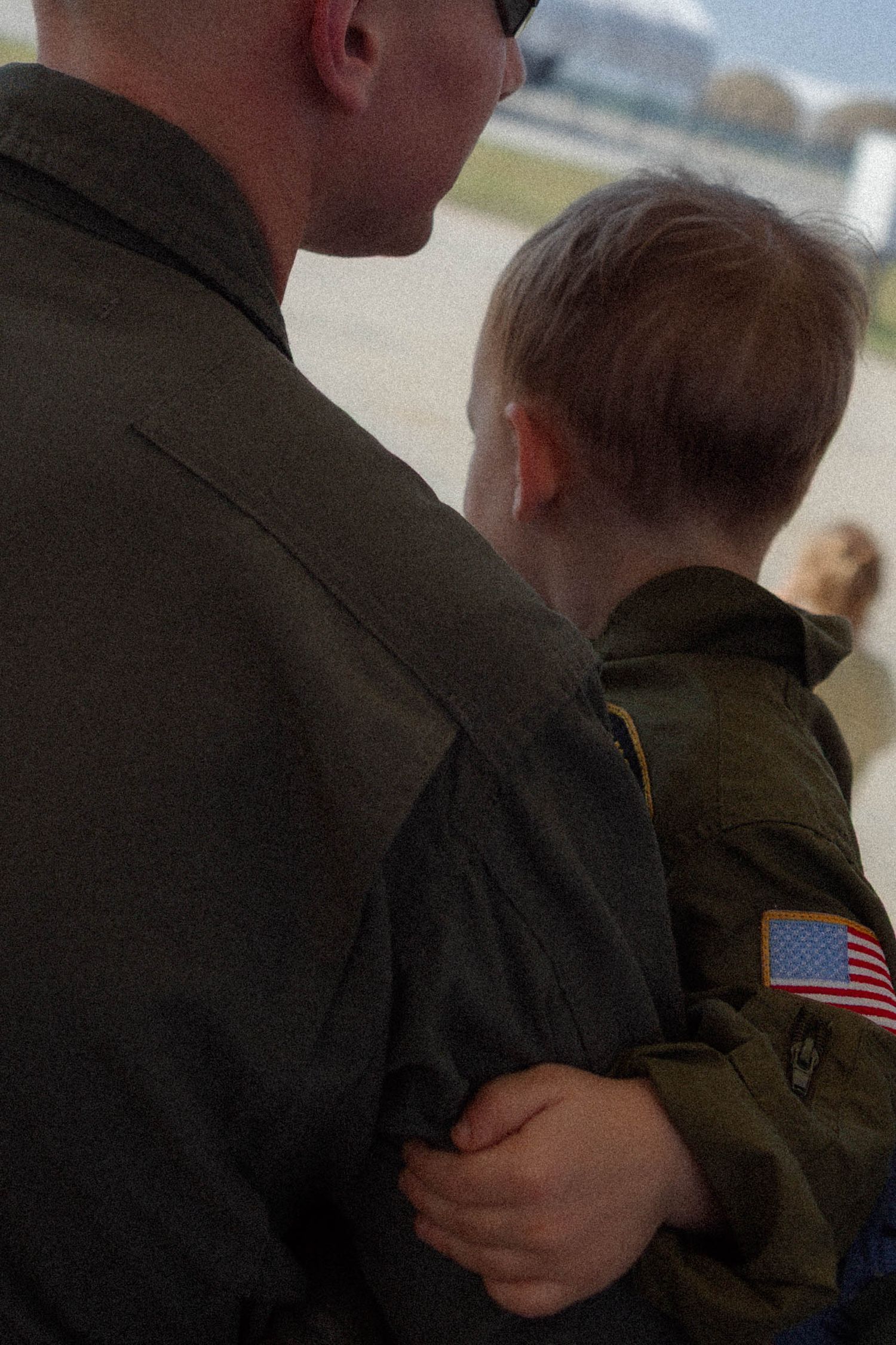A military father in flight suit holds his young son dressed in matching uniform on an airfield.