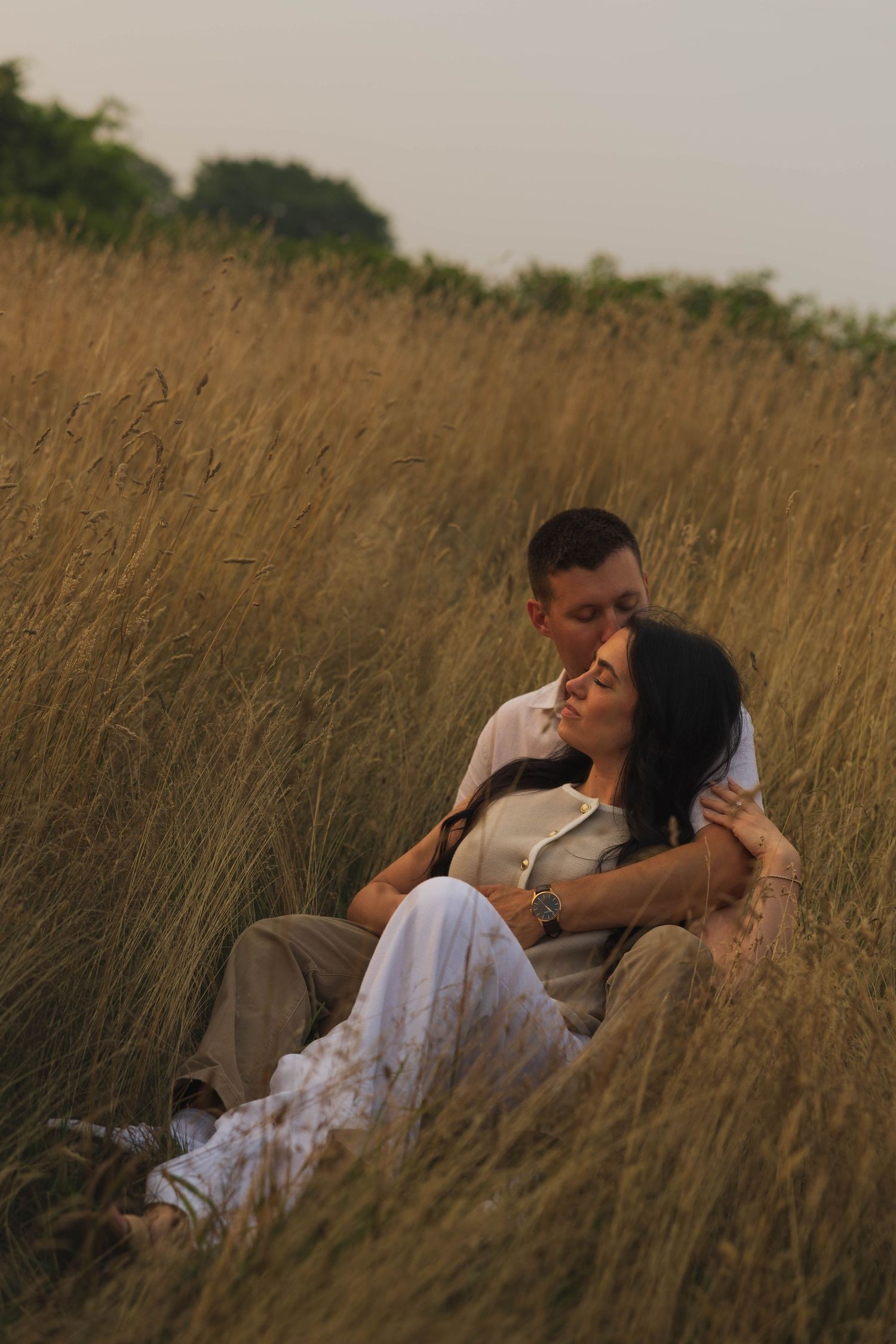 A romantic couple cuddles together sitting in tall golden grass fields at sunset.