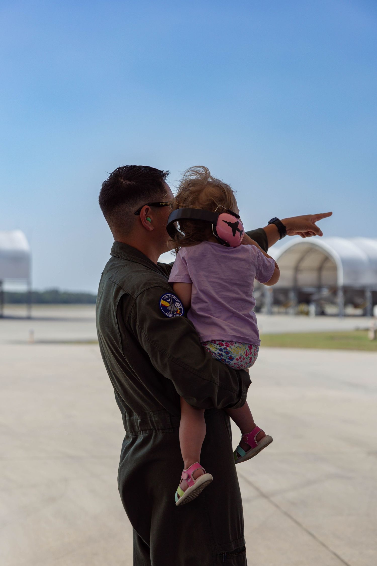 Military pilot in olive flight suit holds toddler girl with pink ear protection on sunny airfield tarmac.