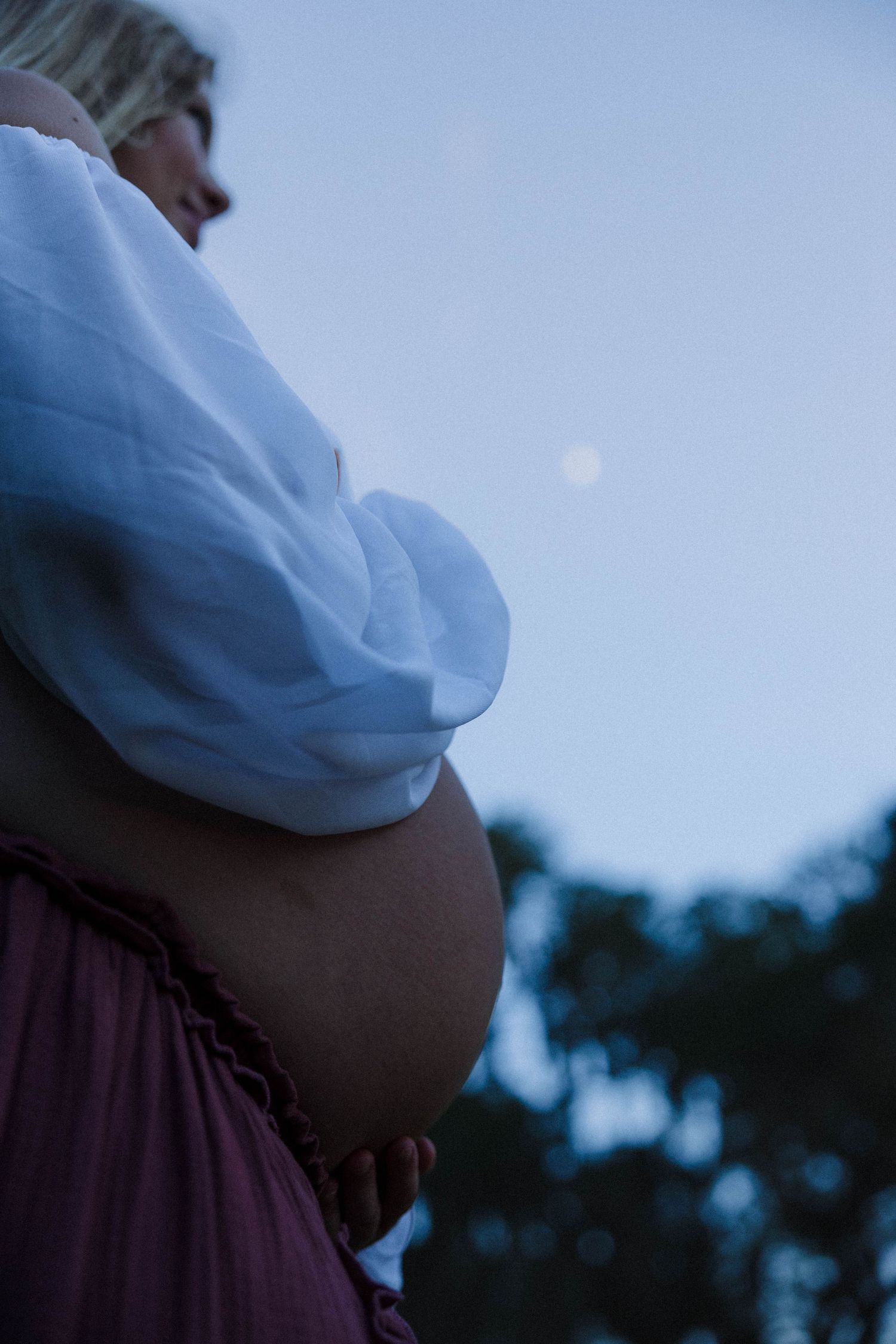Pregnant woman cradles bare baby bump under moonlit twilight sky wearing a flowing white top.