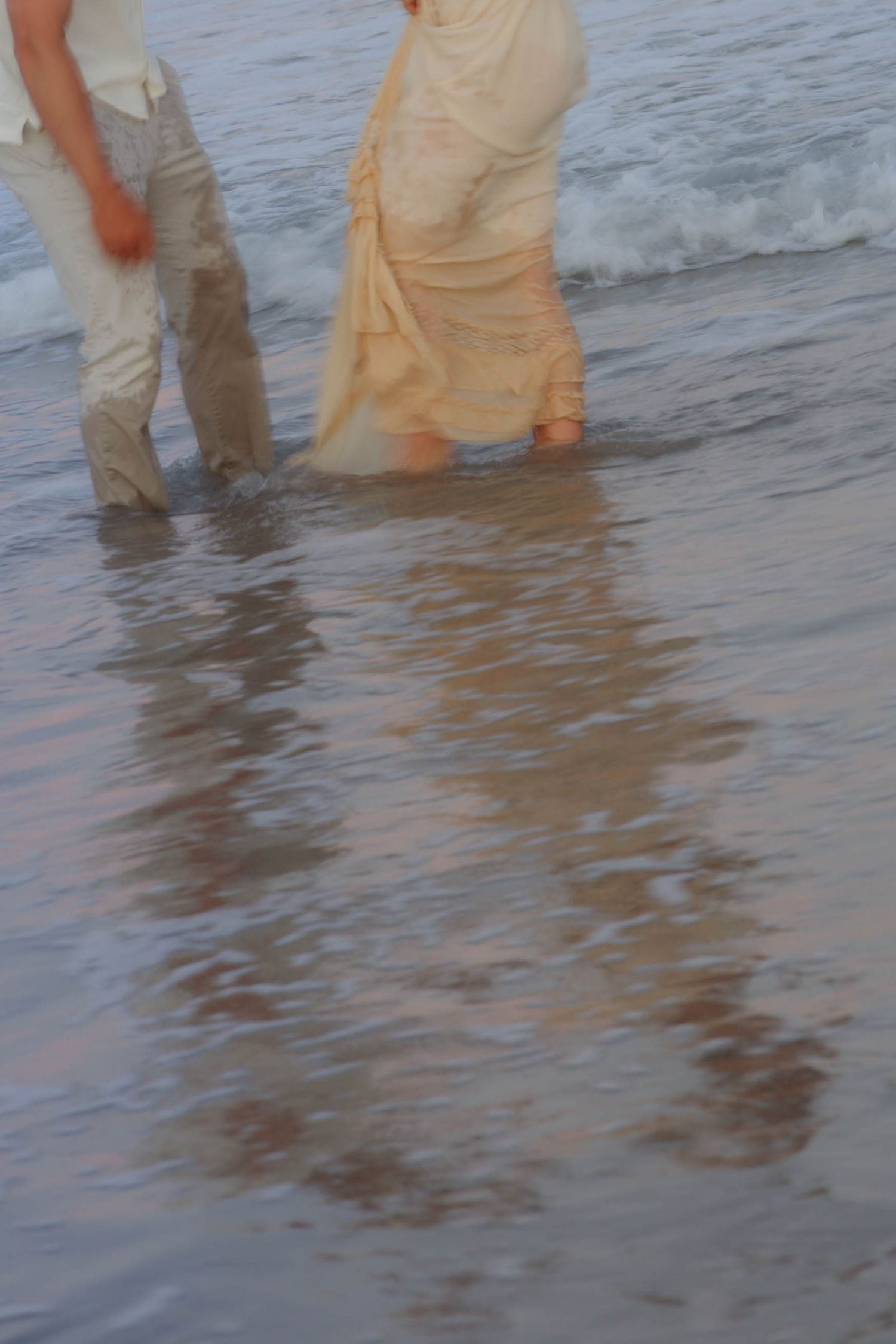 Blurred motion shot of couple wading in ocean surf, bride in white dress and groom in light pants.