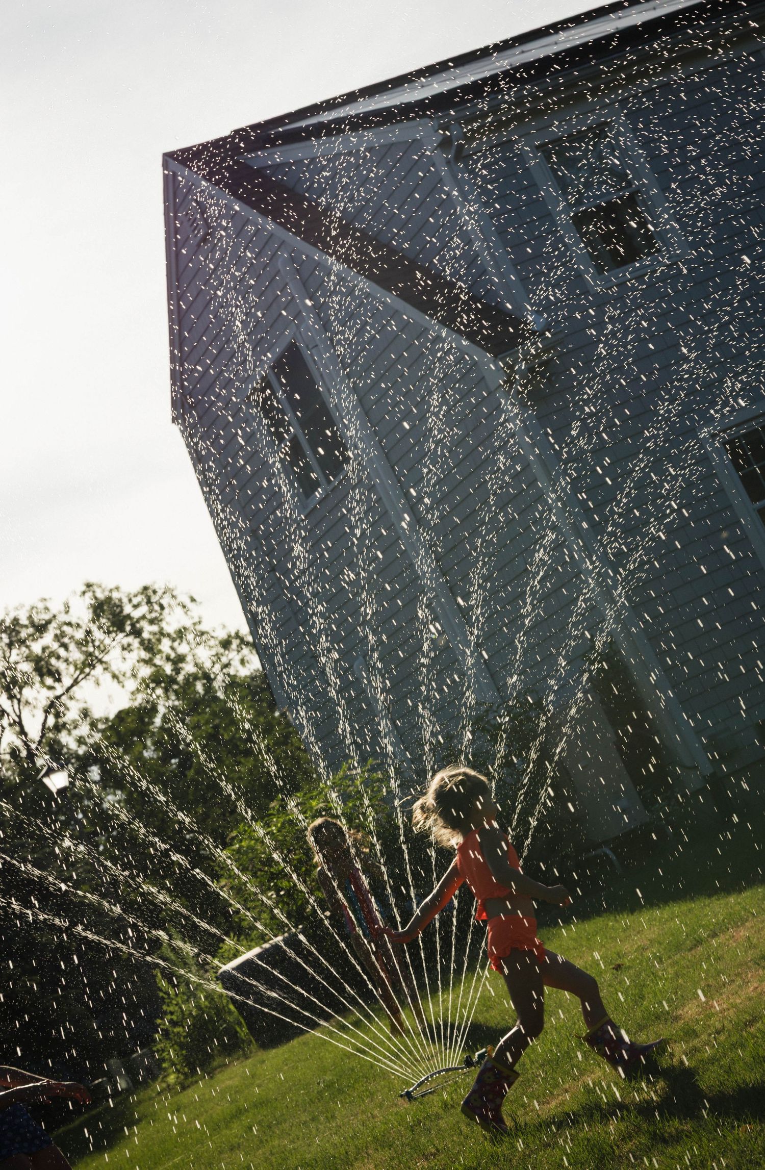 A child runs joyfully through a backlit garden sprinkler creating a stunning water arc display.
