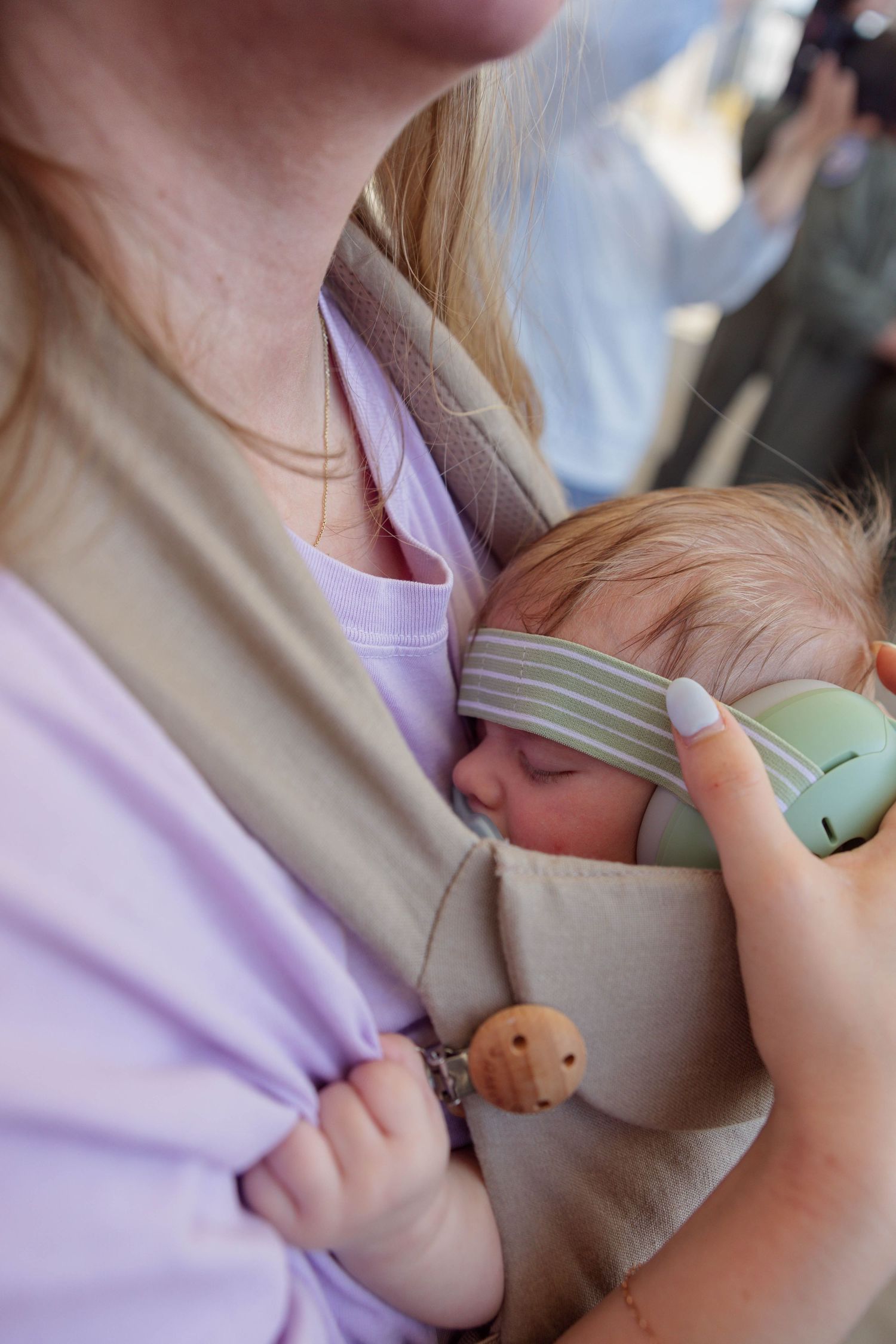A sleeping infant in a baby carrier holds a toy phone while being carried by a blonde mother.