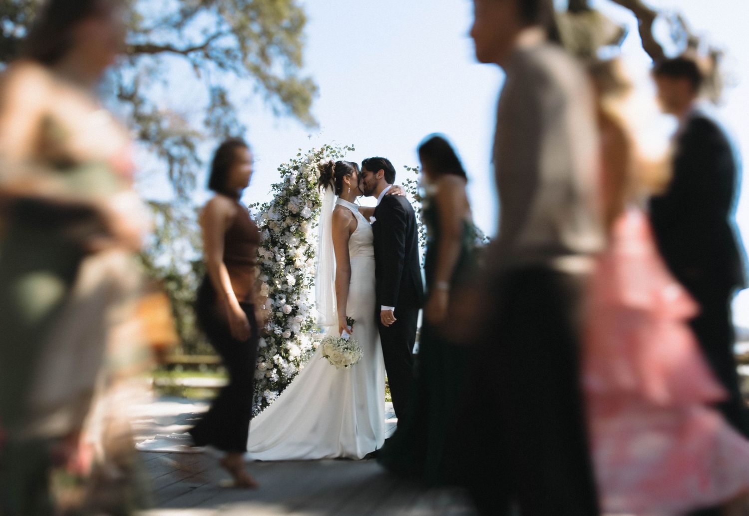 A bride and groom share a first kiss at a floral arch altar surrounded by wedding guests in soft focus.