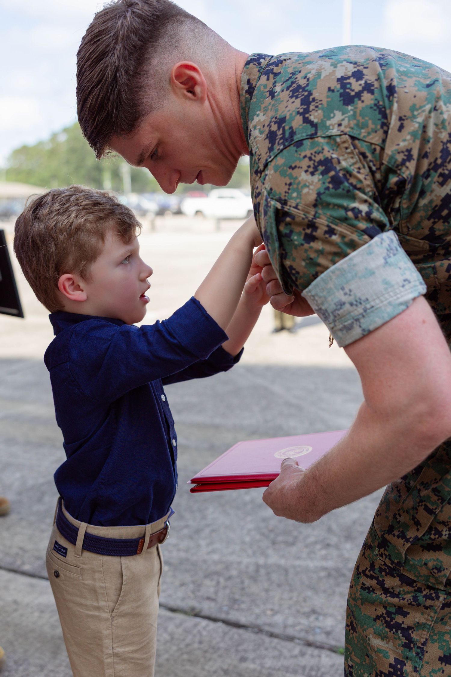 A Marine in camouflage uniform bends down to interact with a young boy in a blue shirt during a military ceremony.