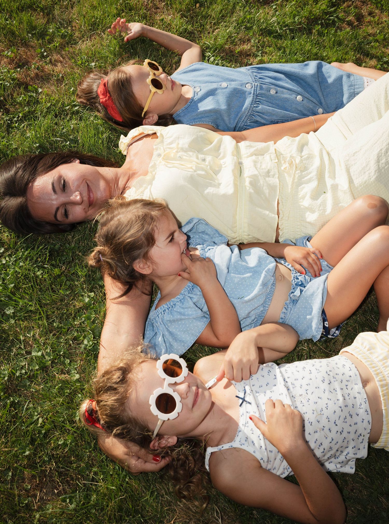 A mother and three young daughters lie together on green grass wearing sunglasses on a sunny day.
