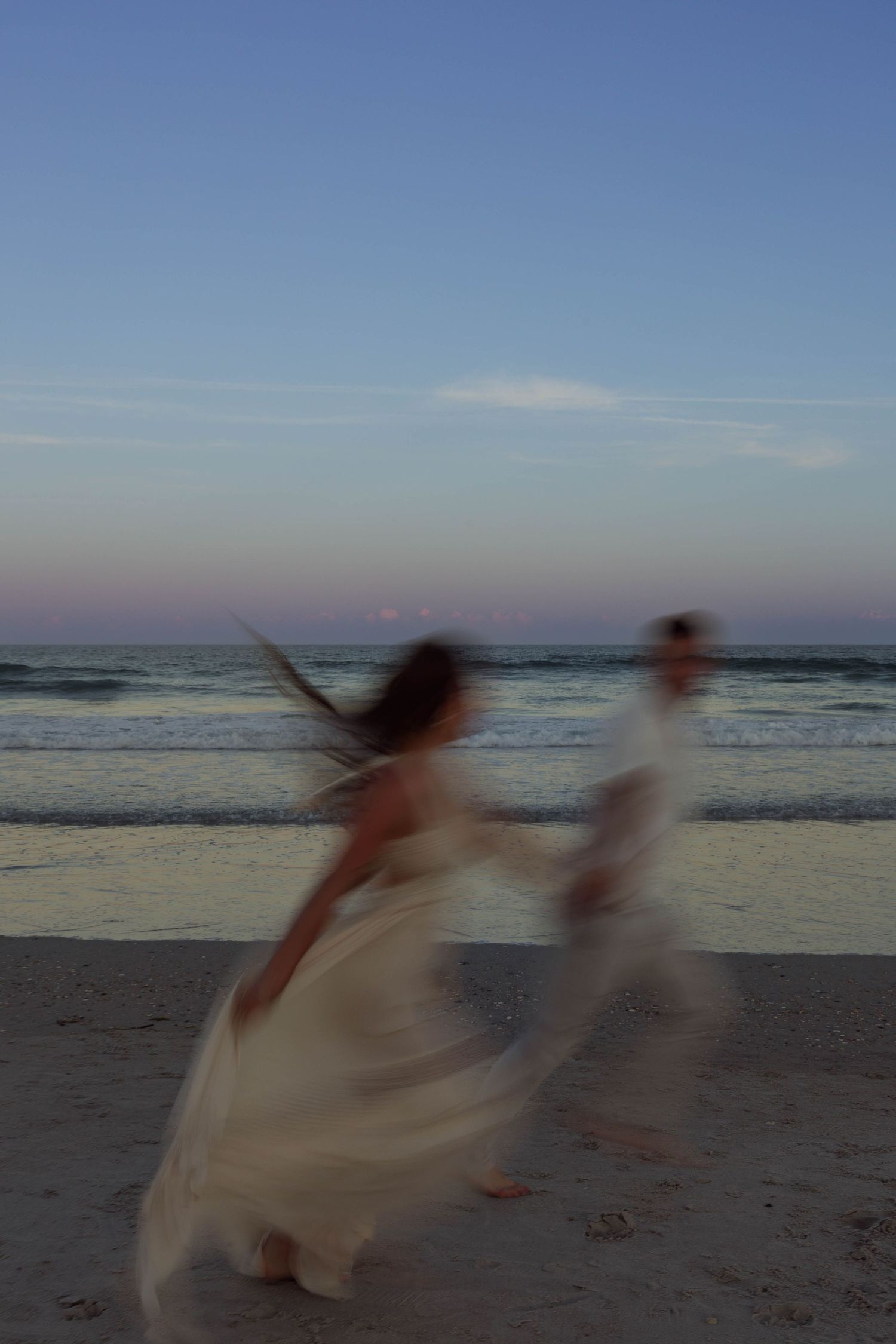 A blurred couple in wedding attire dances romantically on a beach at sunset with ocean waves behind them.