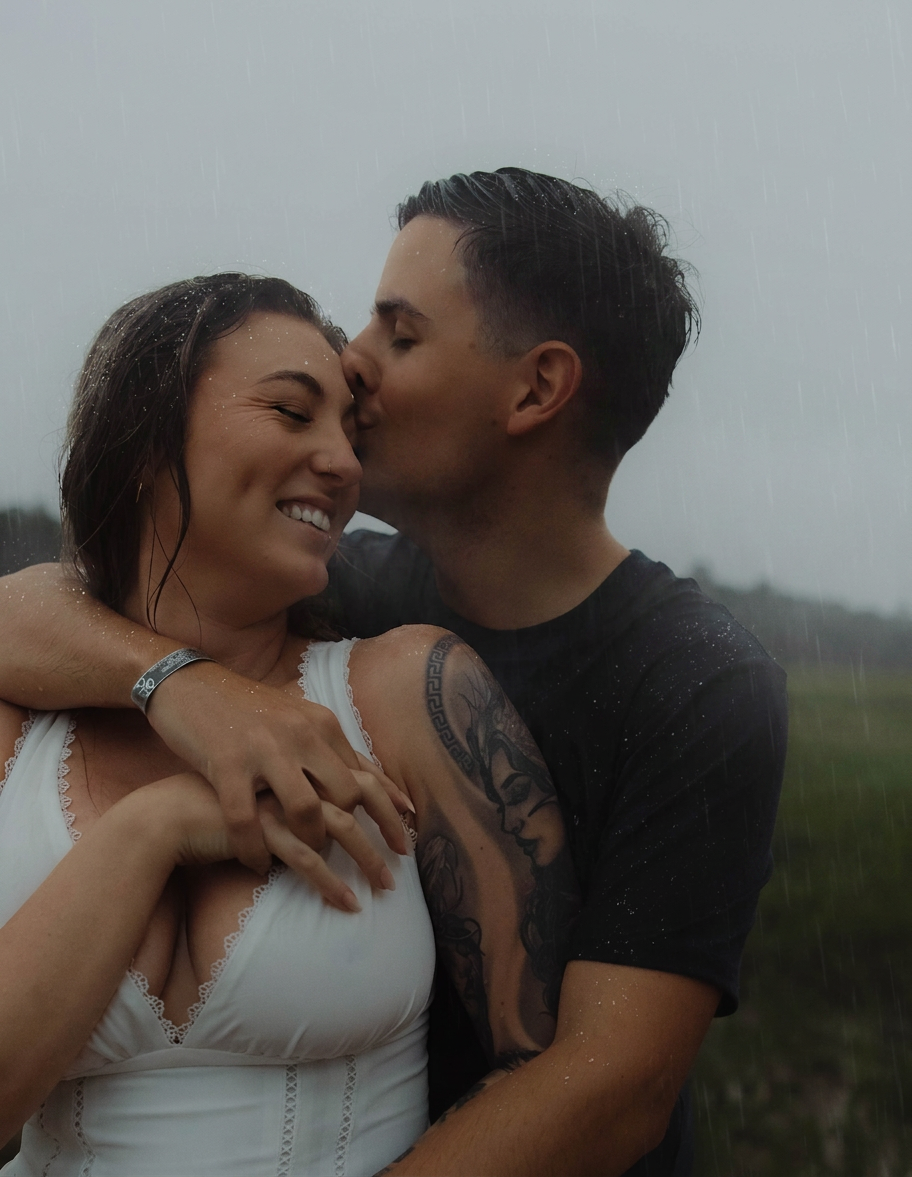 A tattooed man in a black shirt kisses a smiling woman in white on a misty outdoor hillside.