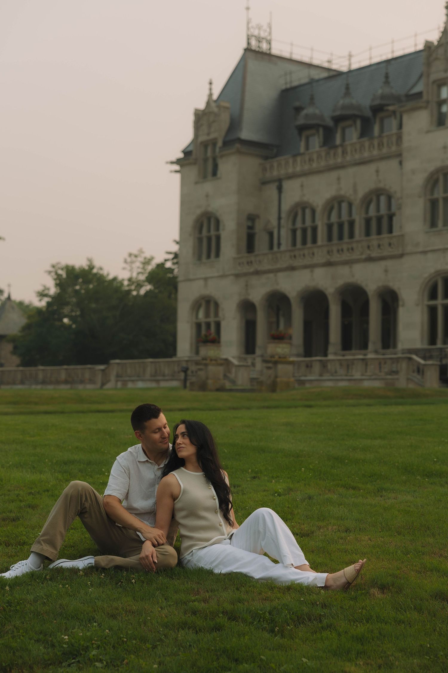 A couple sits together on green grass in front of a grand historic stone mansion at dusk.