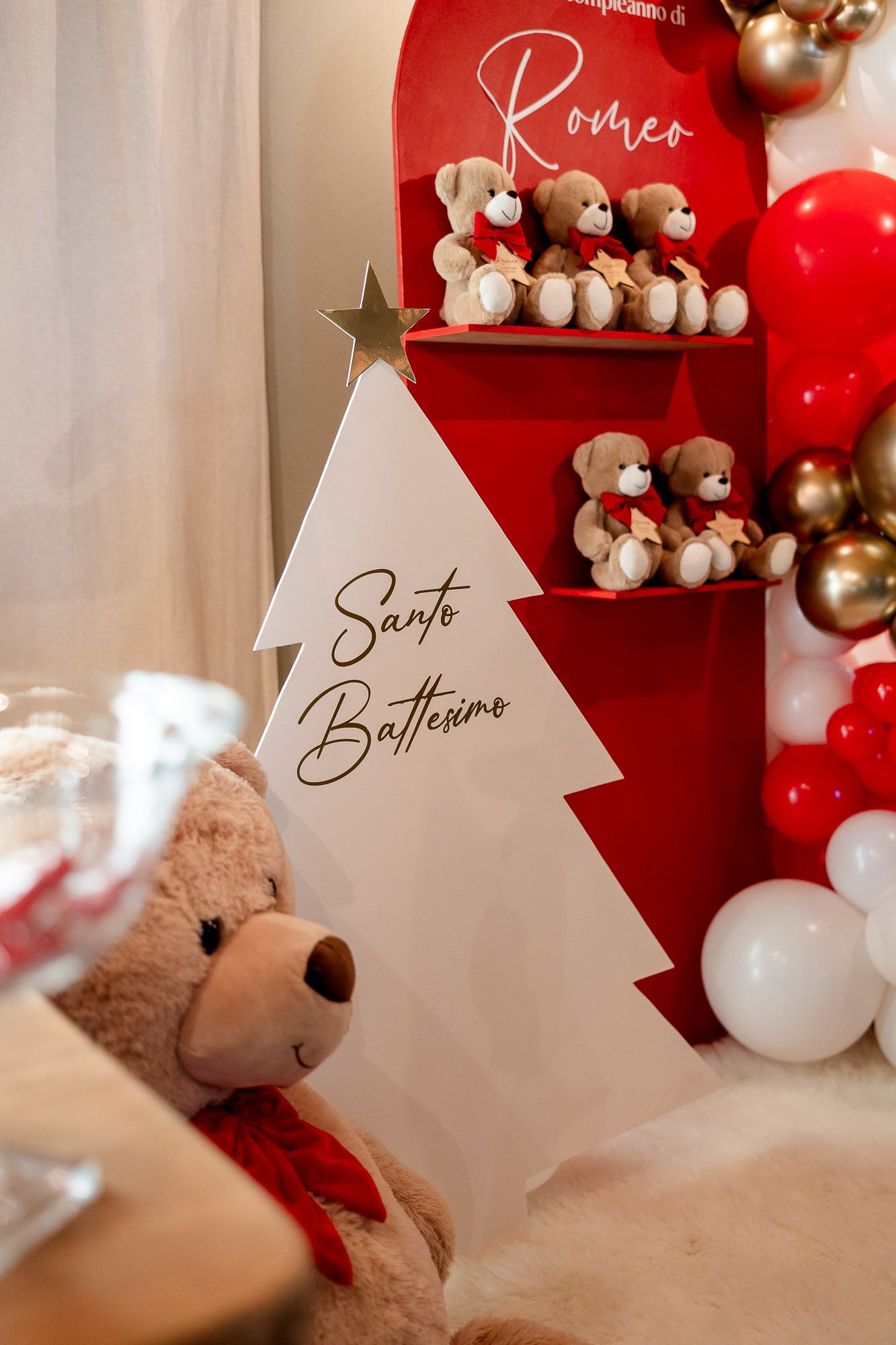 Red and white baptism party decor with teddy bears, balloons, and a tree sign reading Santo Battesimo for Romeo.