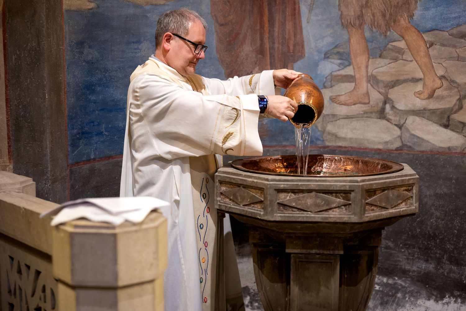 Priest in white vestments pouring water into a copper baptismal font during a church baptism ceremony.