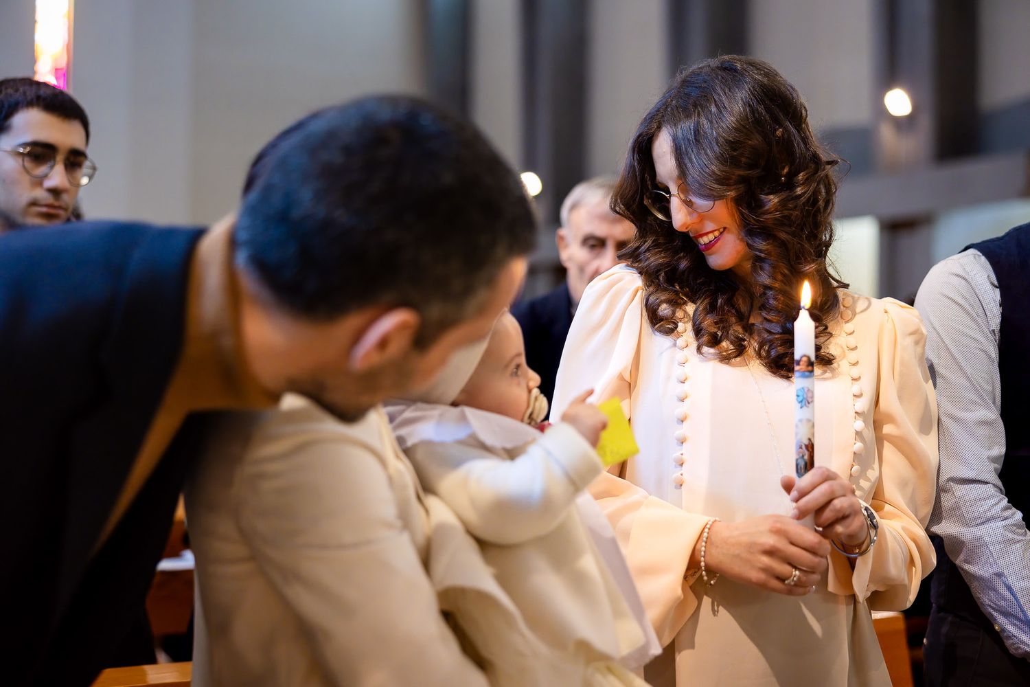 A woman in white holds a candle during a baptism ceremony in a church, smiling at a baby held by a man.