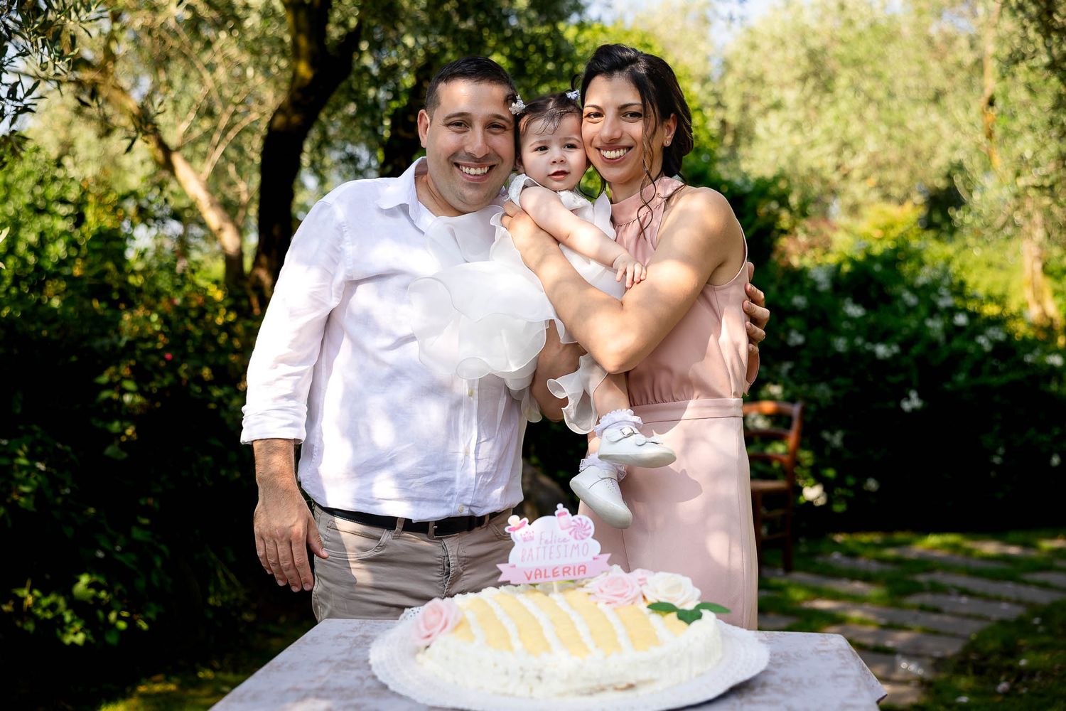 Family celebrating baby first birthday outdoors with a decorated cake in a lush garden setting.