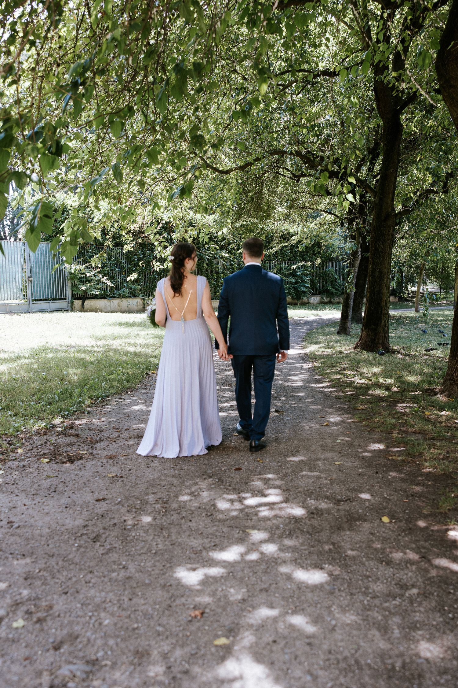 A couple in wedding attire walks down a shaded dirt path surrounded by lush green trees on a sunny day.