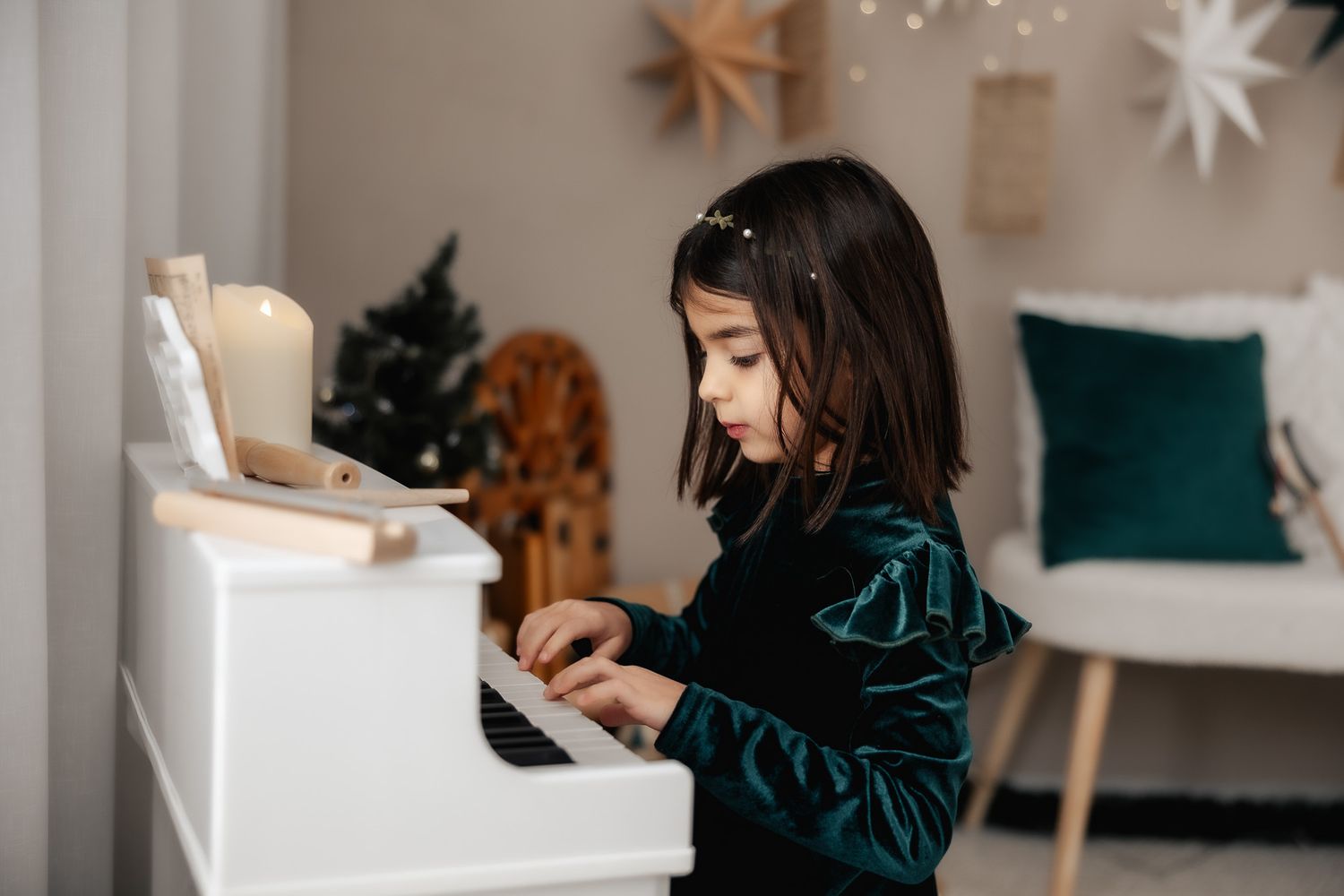 Young girl in green velvet dress playing white toy piano near Christmas tree in cozy decorated room.