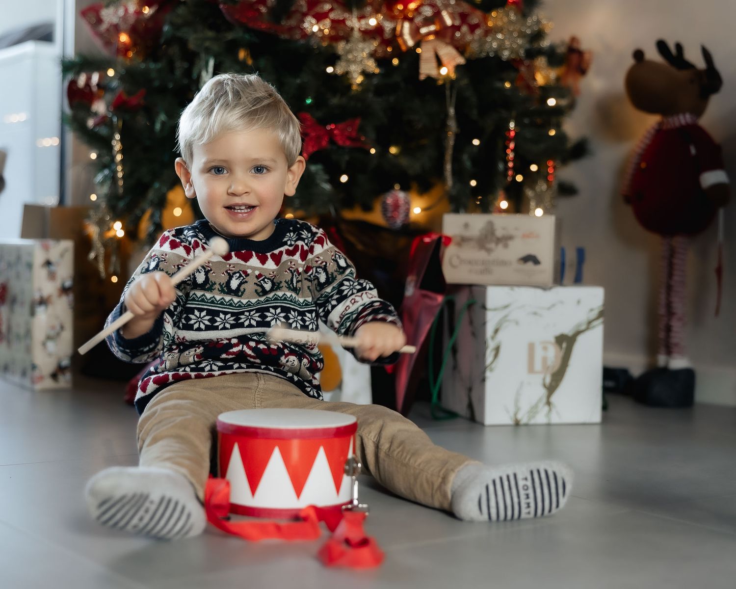 Toddler boy in festive sweater playing with toy drum beside decorated Christmas tree and wrapped gifts.