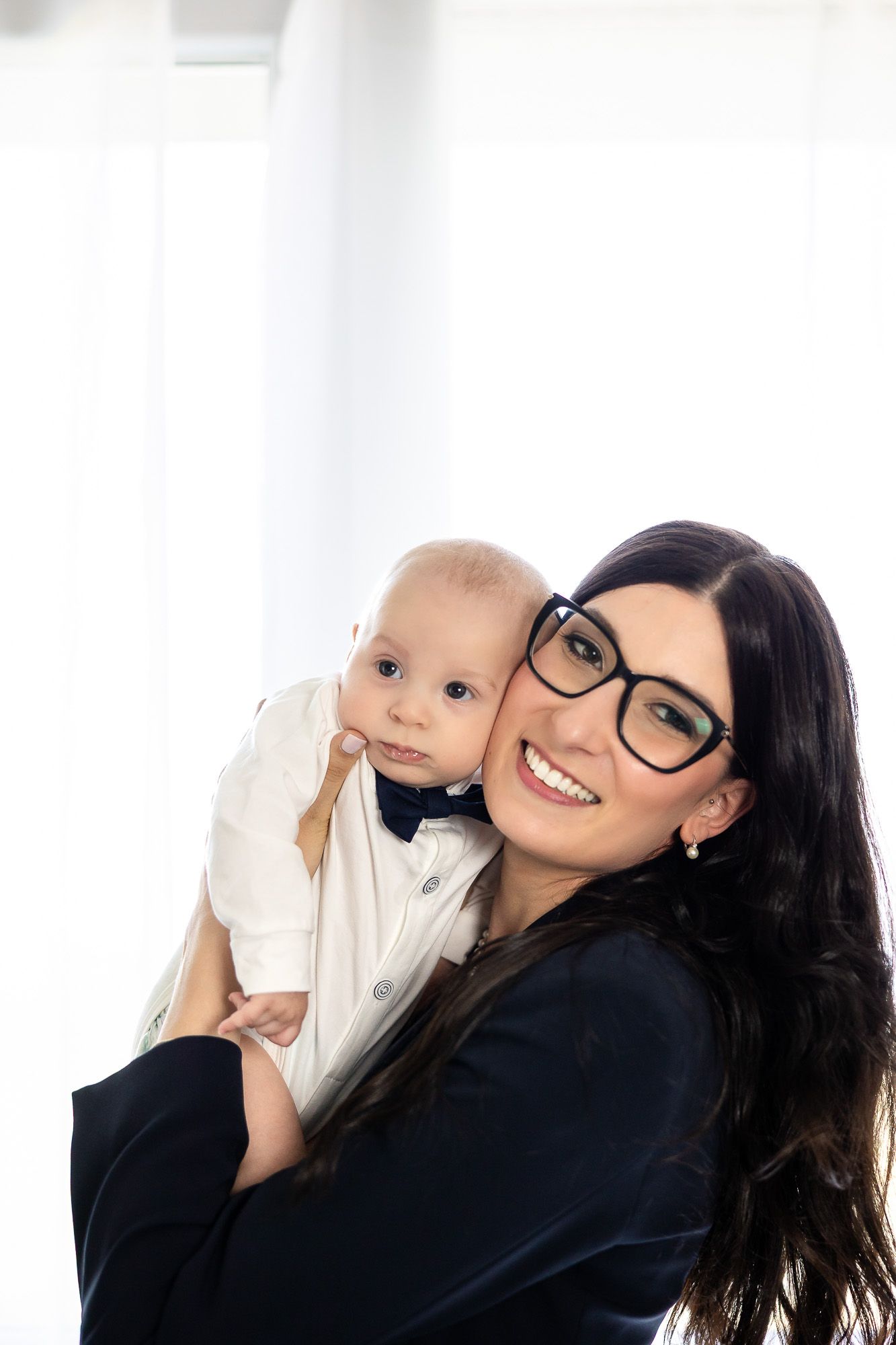Smiling brunette mother with glasses holding her baby boy dressed in white shirt and bow tie.