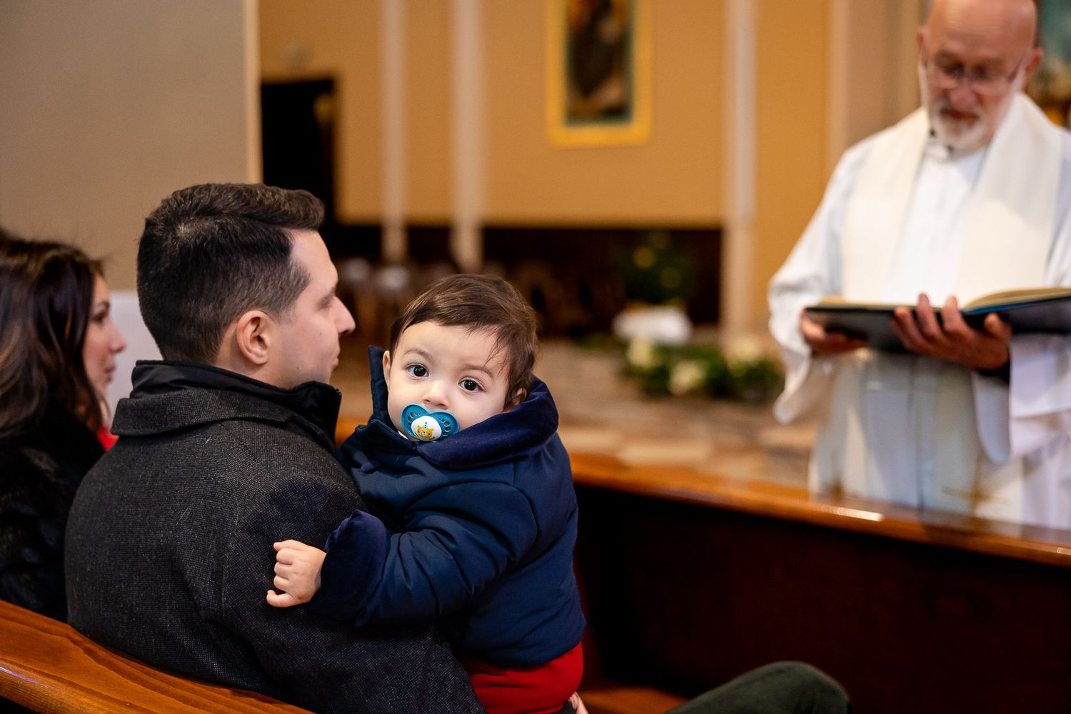 A priest reads from a book during a baptism ceremony as a father holds his baby with a pacifier in a church.