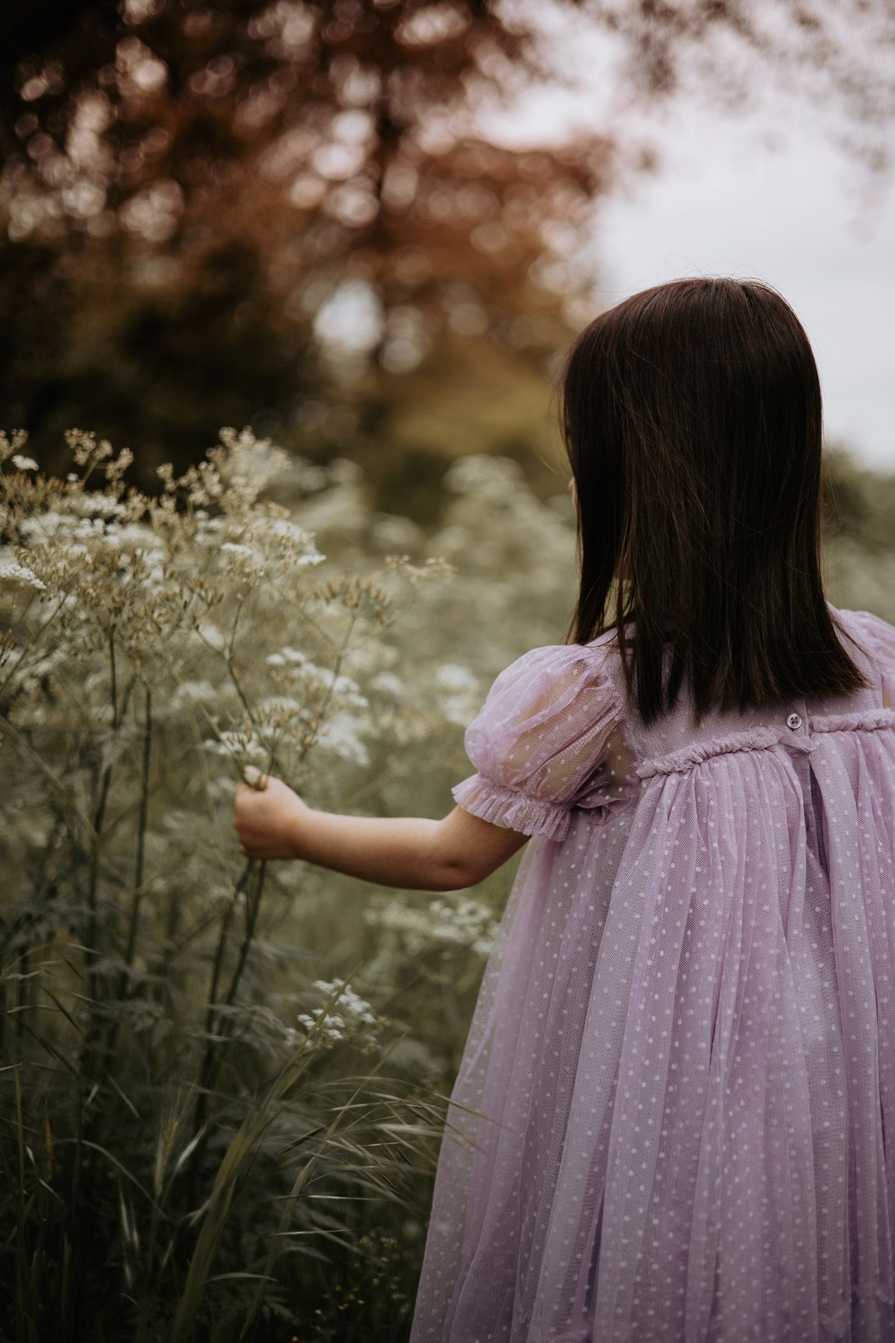 A child in a lavender dress touching wildflowers in a dreamy outdoor setting.