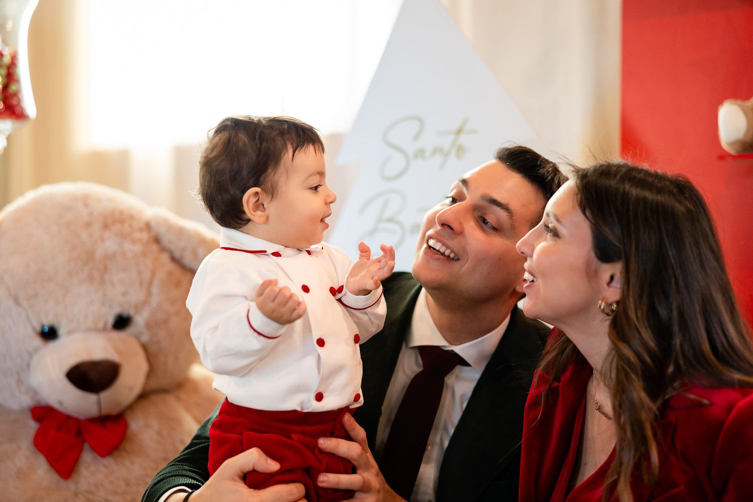 Family with baby celebrating Christmas, parents smiling at infant in festive outfits near large teddy bear and Santa sign.