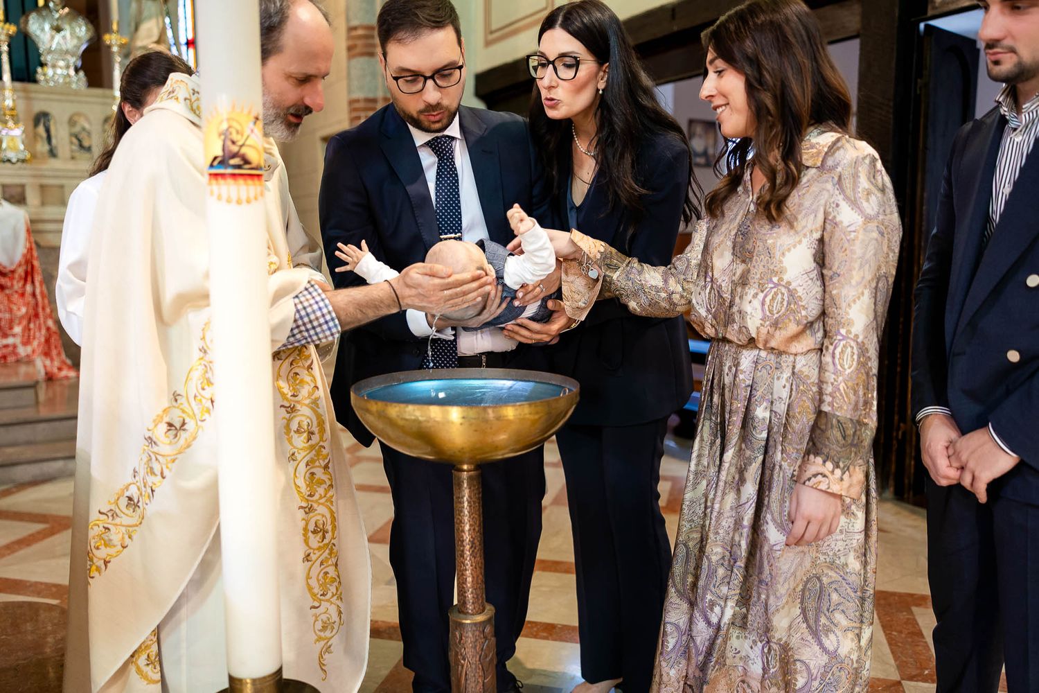 Priest performing Catholic baptism ceremony with family gathered around ornate gold baptismal font in church.