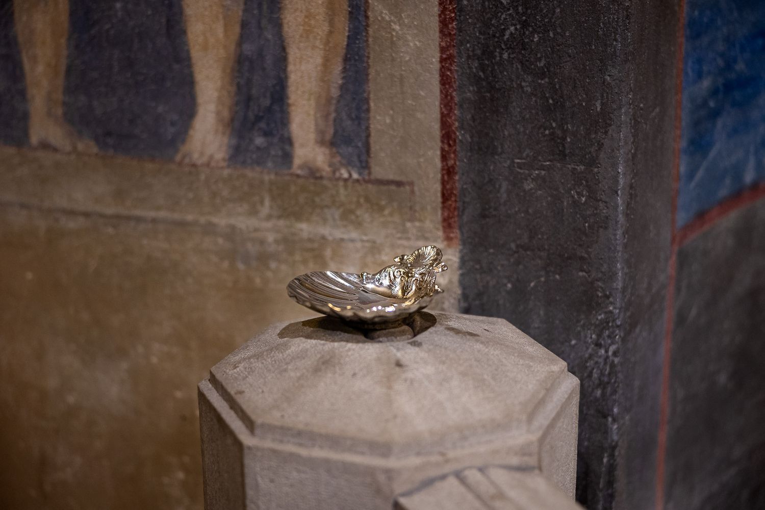 Silver shell-shaped holy water font on stone pedestal in historic church with colorful frescoes.