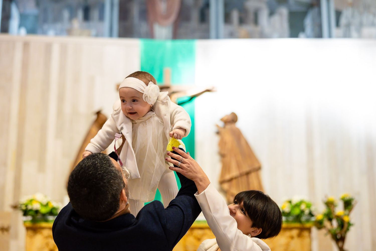 Parents joyfully lift their baby in white baptism outfit at a church altar during a christening ceremony.