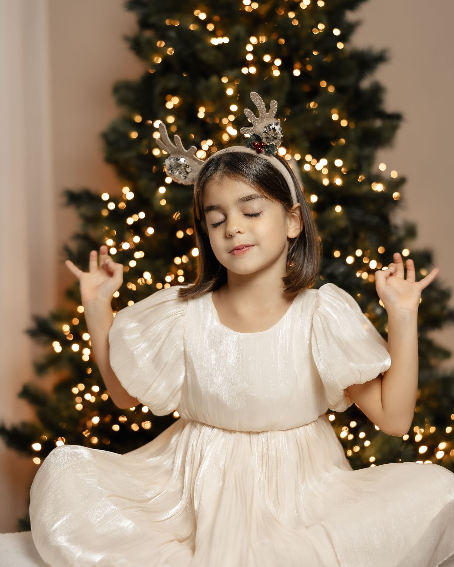 Young child in white dress poses playfully in front of illuminated Christmas tree with twinkling lights and decorations.