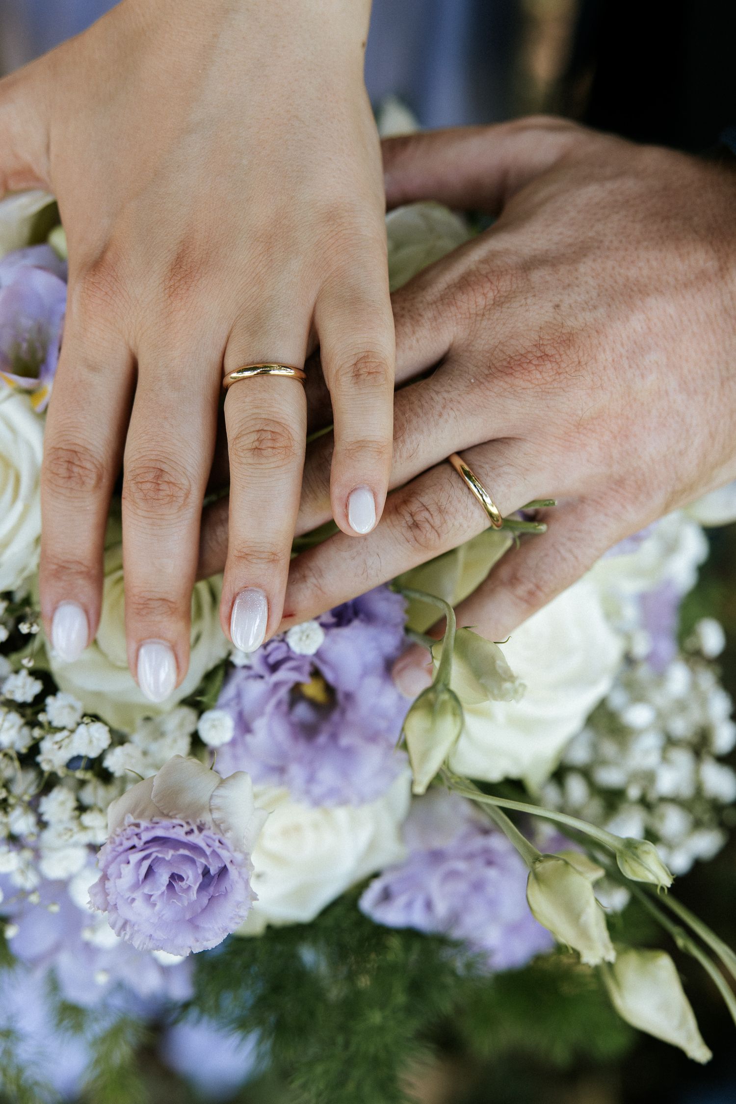 Close-up of hands holding delicate purple and white flowers with wedding rings visible.
