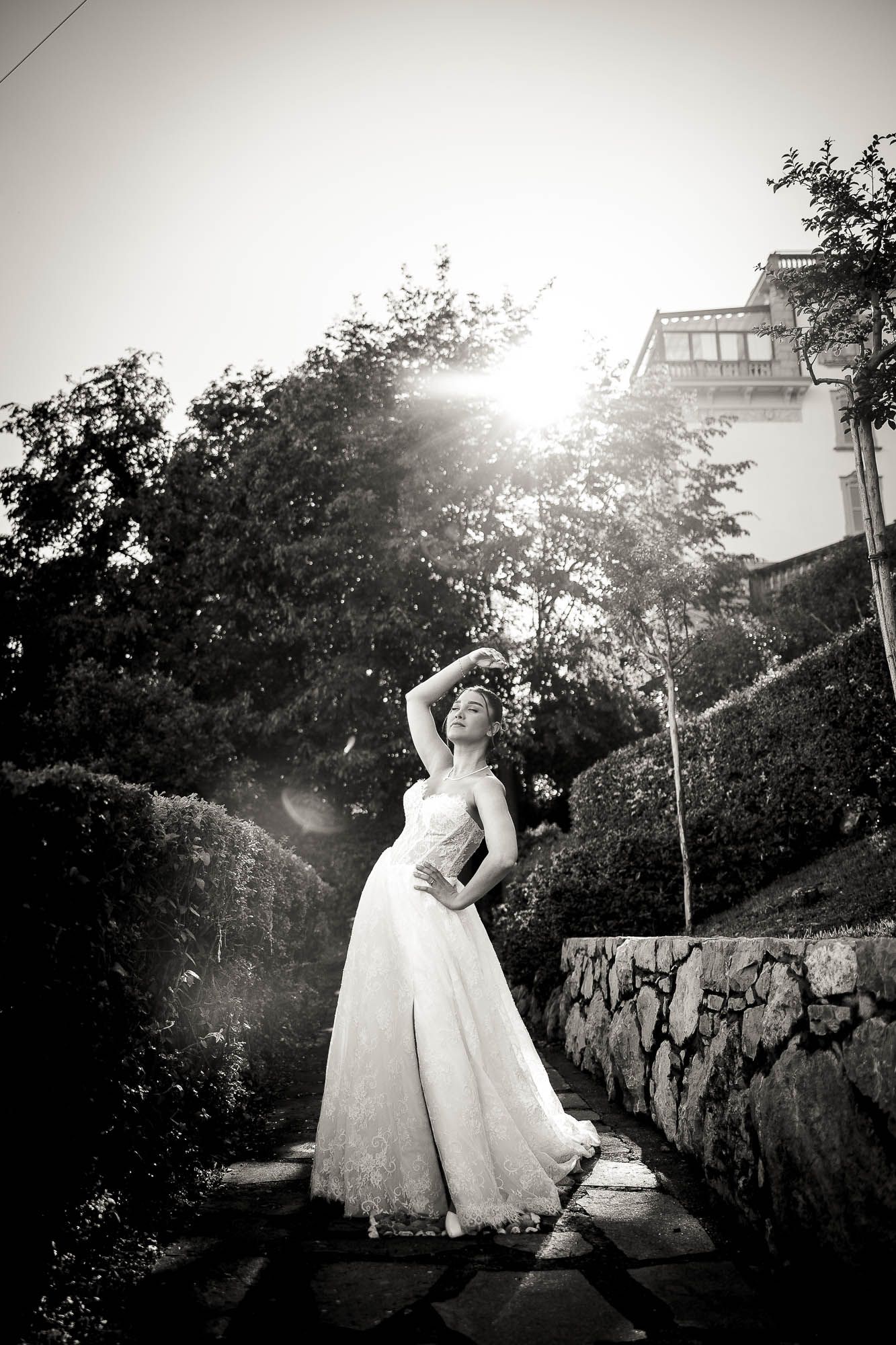 Bride in white lace gown poses gracefully outdoors in black and white photo with sunlight and stone wall backdrop.