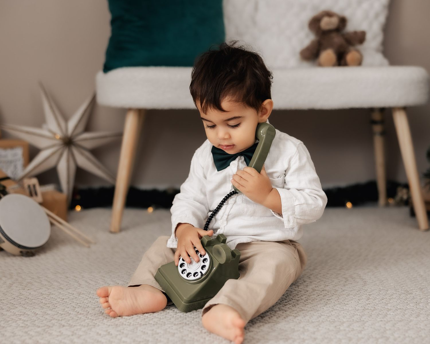 A cute toddler boy in a white shirt and bow tie sits on a floor, pretending to talk on a vintage rotary phone.