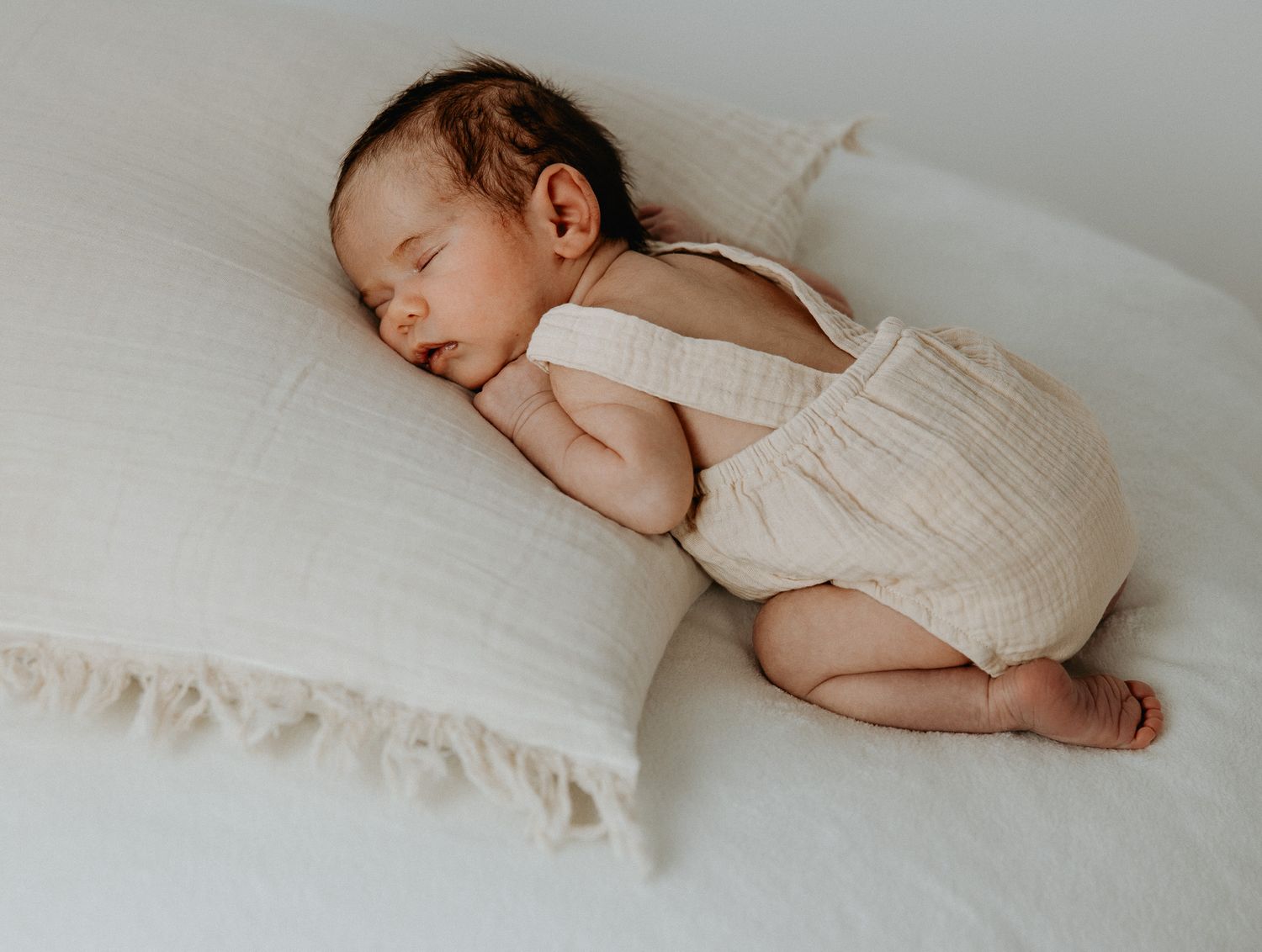 A sleeping baby wearing white clothing lies peacefully on soft white bedding.