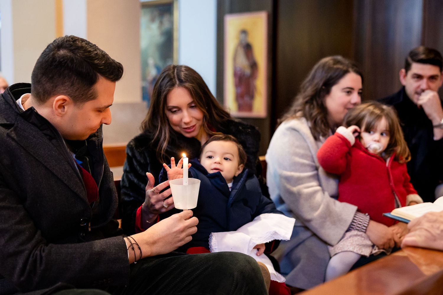 Family gathered indoors, parents smiling at baby holding a cup during a christening ceremony.