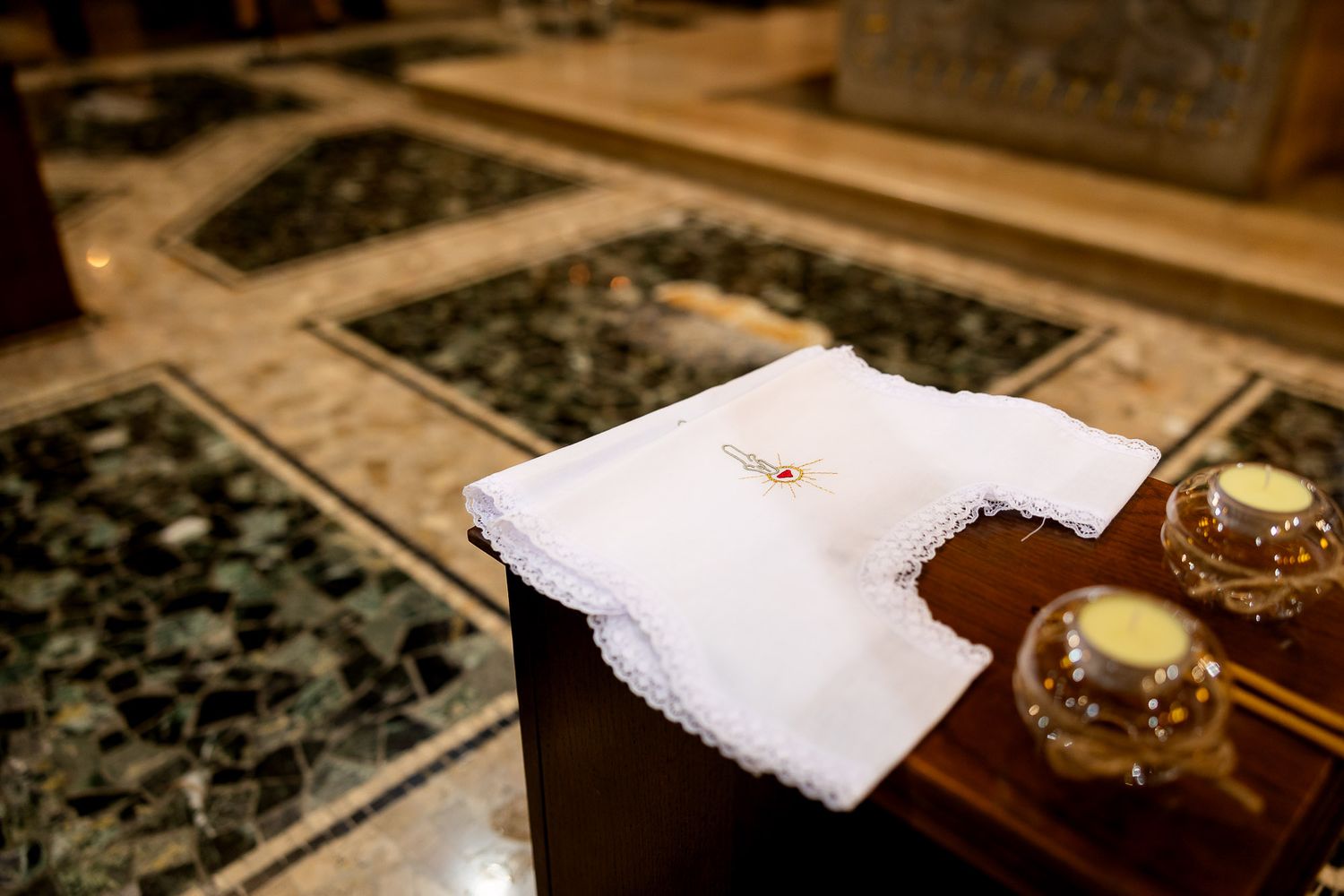 White baptismal cloth with lace trim and gold candles on wooden stand in ornate marble church interior.