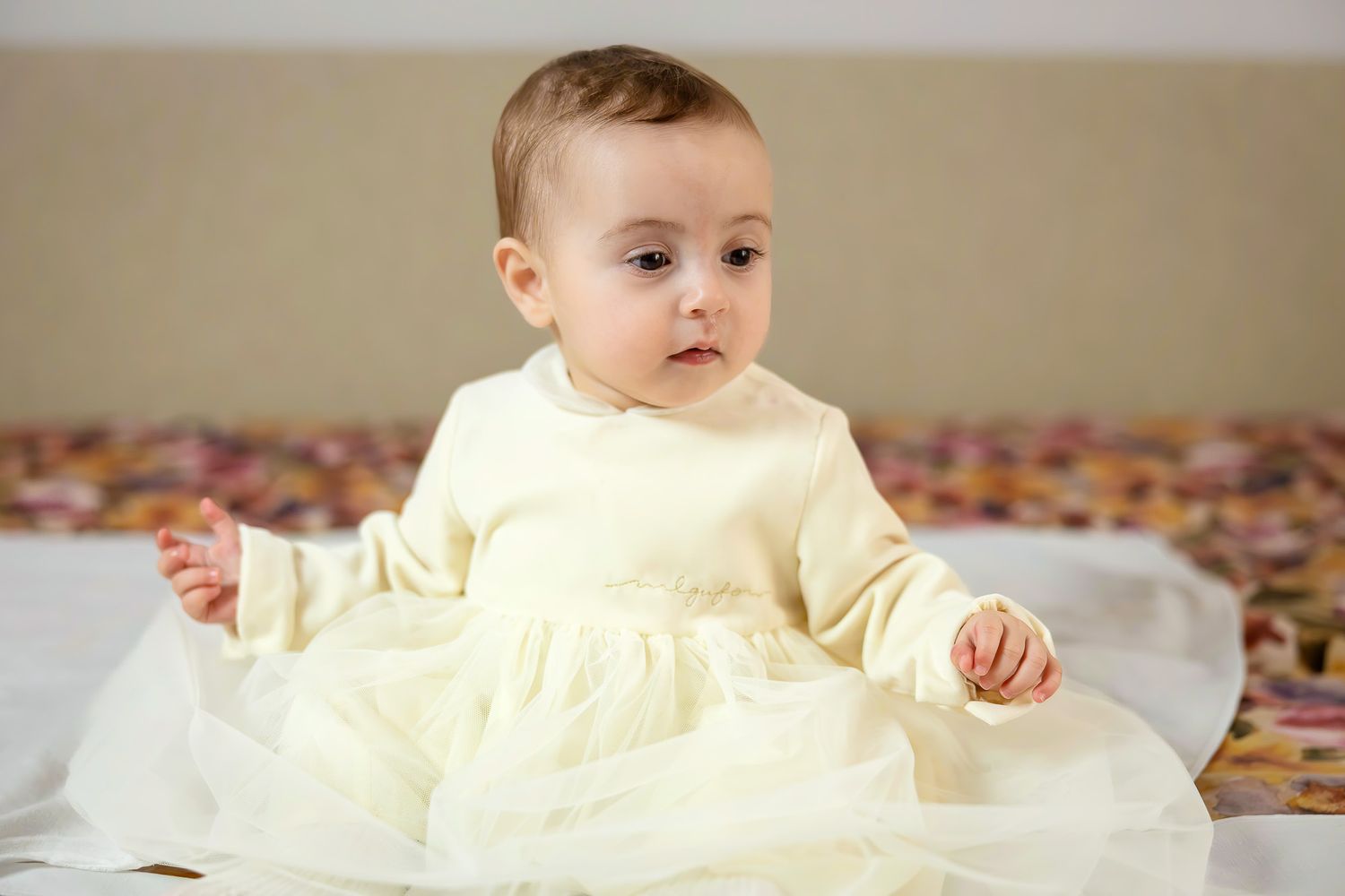 Adorable baby girl in a cream white dress sitting on a bed, looking curiously to the side.