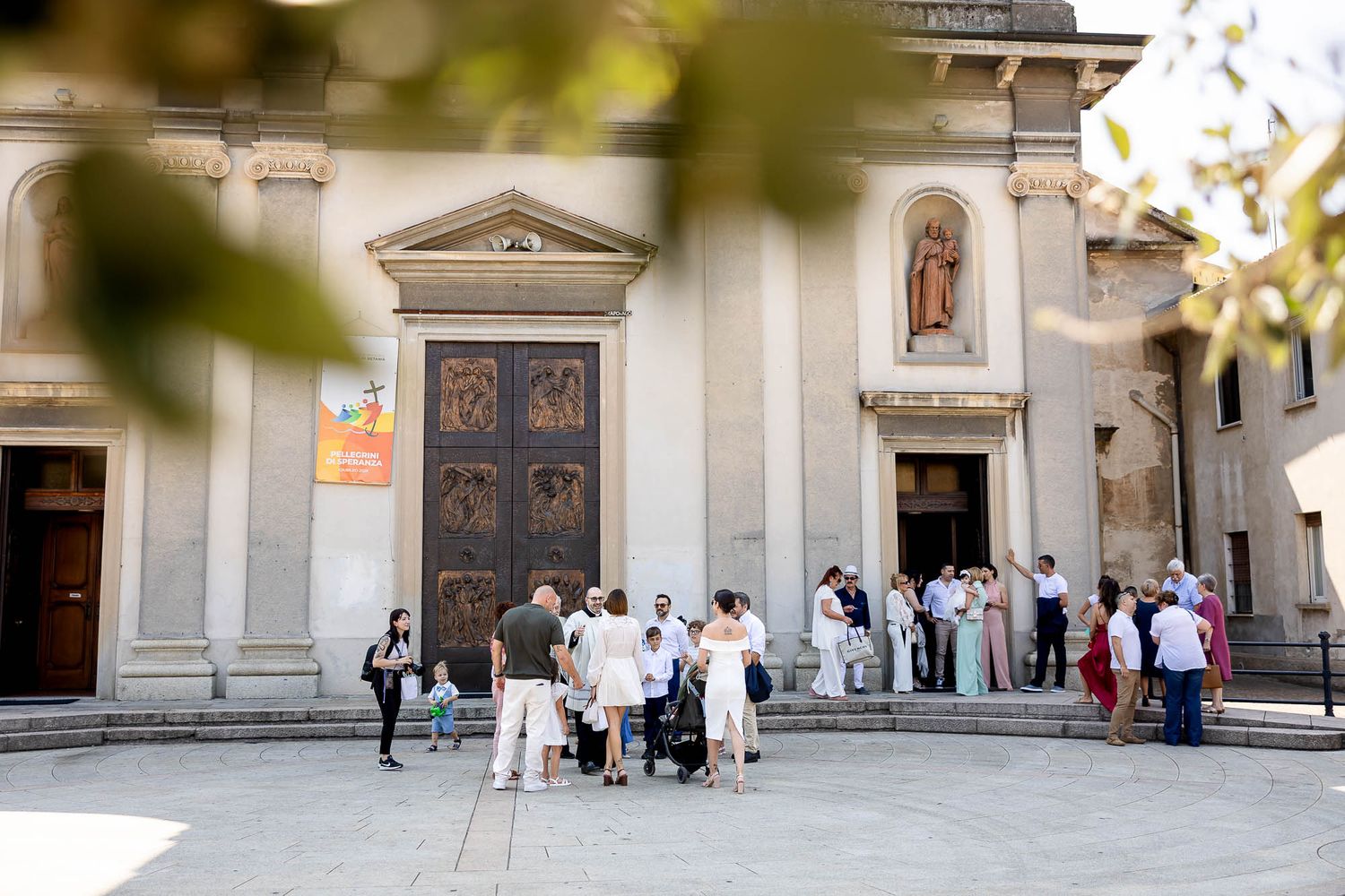 Crowd of people gathered outside a historic Italian church with large wooden doors and classical architecture on a sunny day.