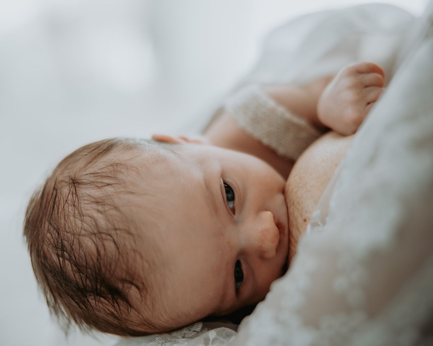 Newborn baby with dark hair lying peacefully in a soft white blanket, gazing gently with bright eyes.