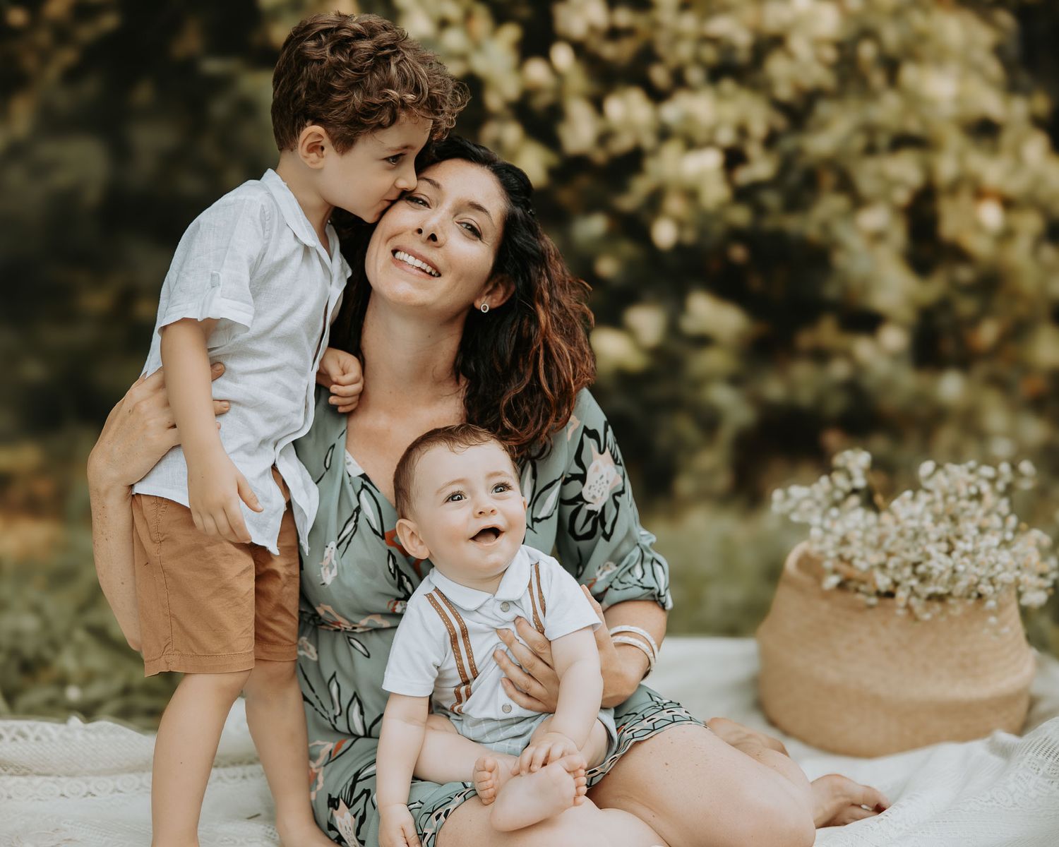 A smiling mother sits outdoors with her two young sons, sharing a joyful embrace surrounded by lush greenery.