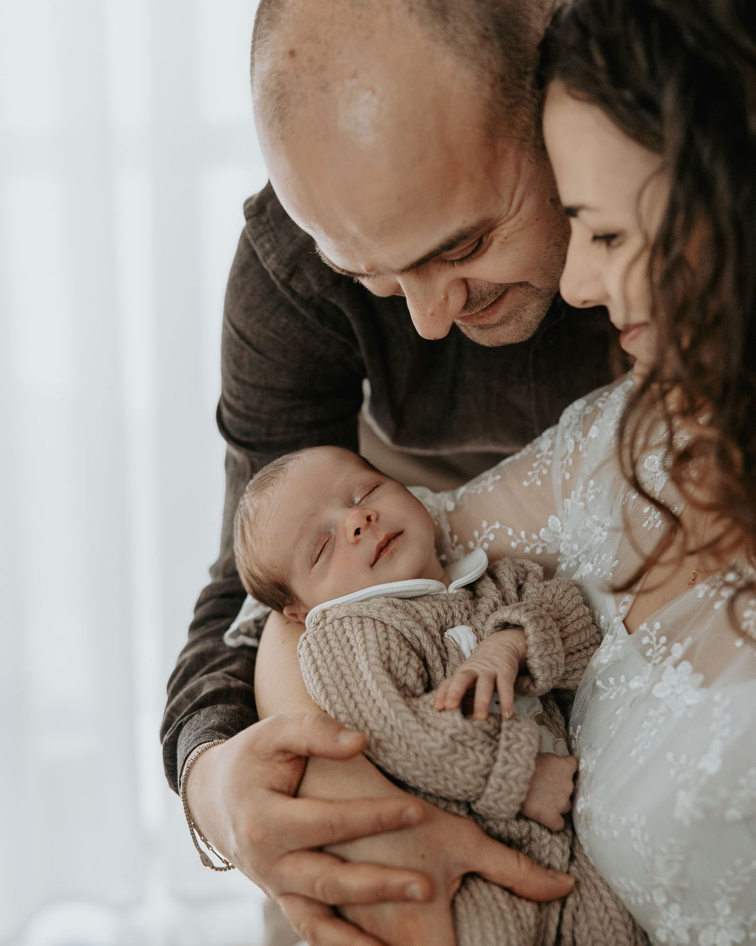 A couple tenderly holds their sleeping newborn baby, sharing an intimate family moment in soft natural light.