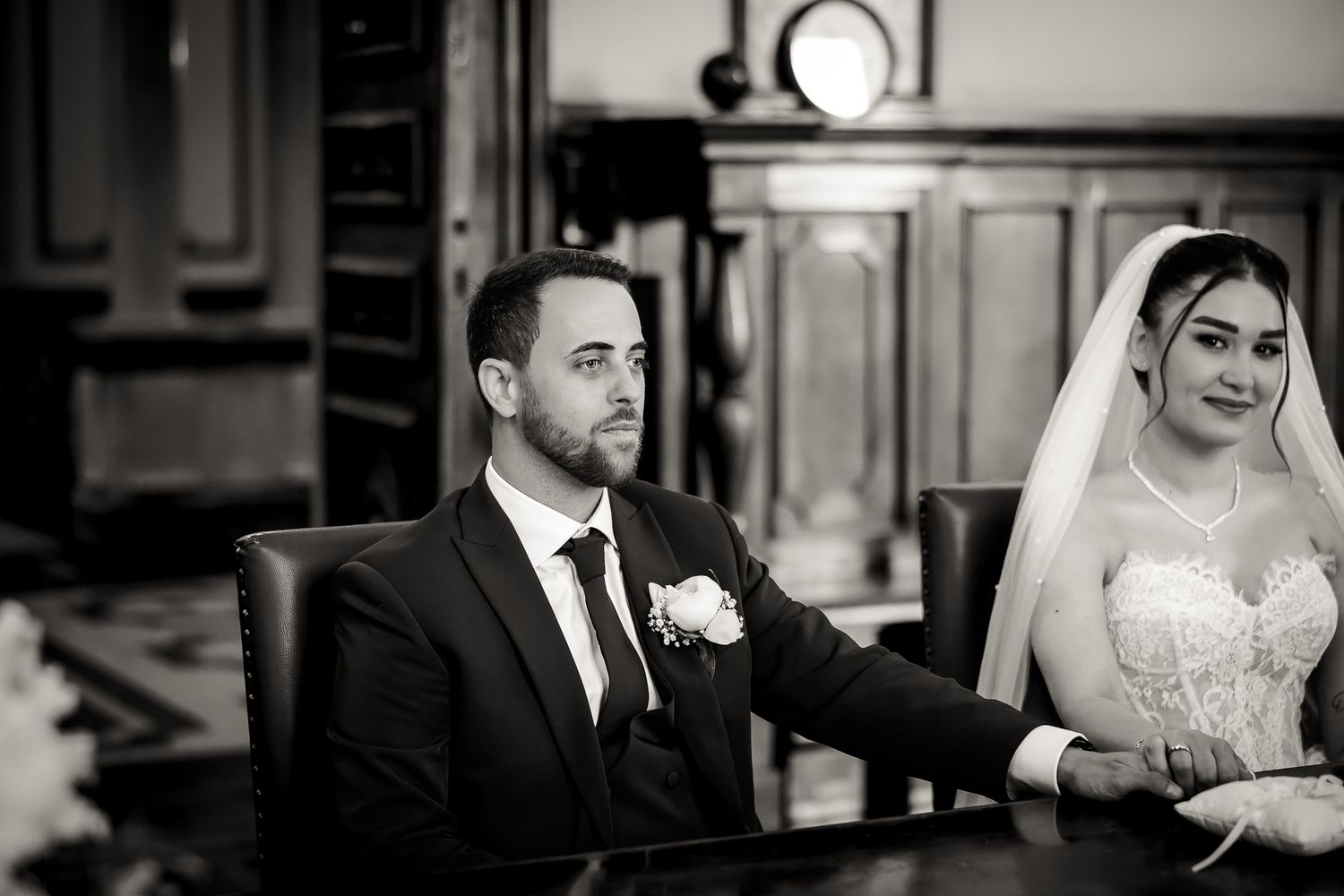Black and white wedding photo of bride and groom seated at a table, bride in veil touching groom's arm.