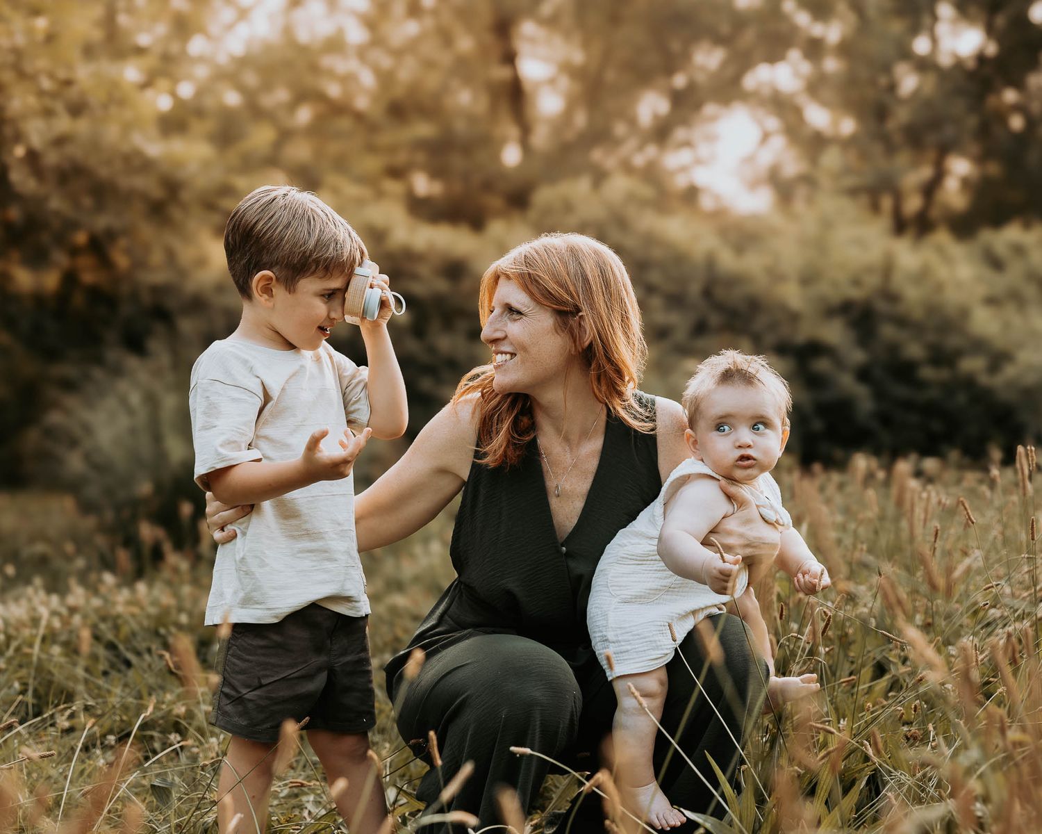 A redheaded woman crouches in a field holding a baby while a young boy photographs her with a camera.