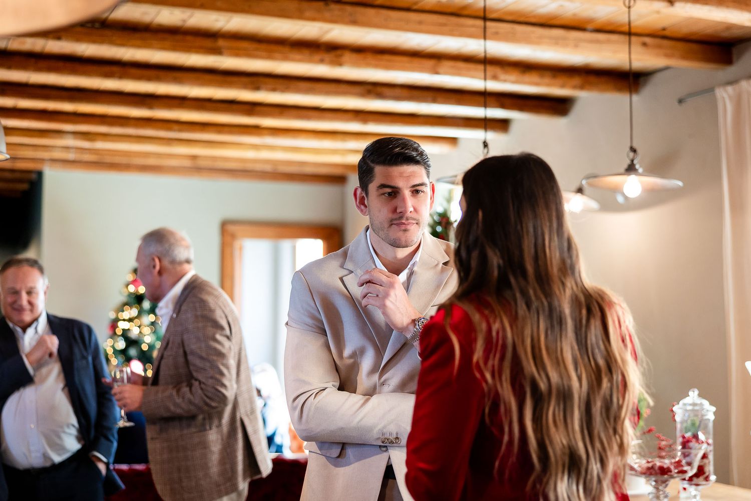 Man in beige blazer adjusting tie while talking to woman in red at an indoor holiday gathering.