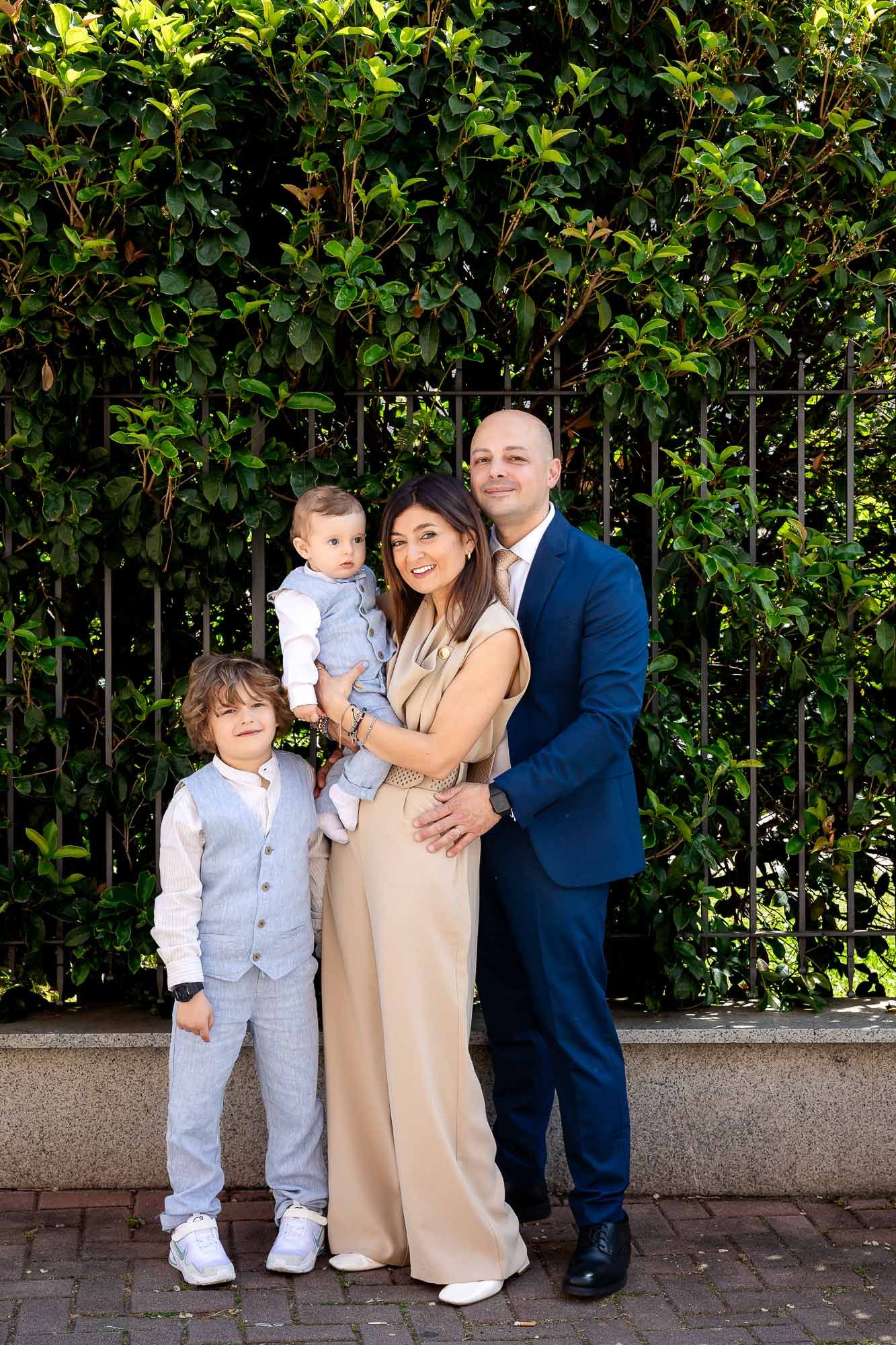Family of four posing outdoors in formal attire, with lush green hedge and iron fence backdrop.