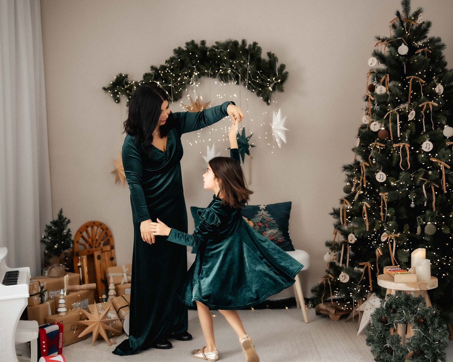 Mother and daughter in matching green velvet dresses decorating for Christmas near a festive tree.
