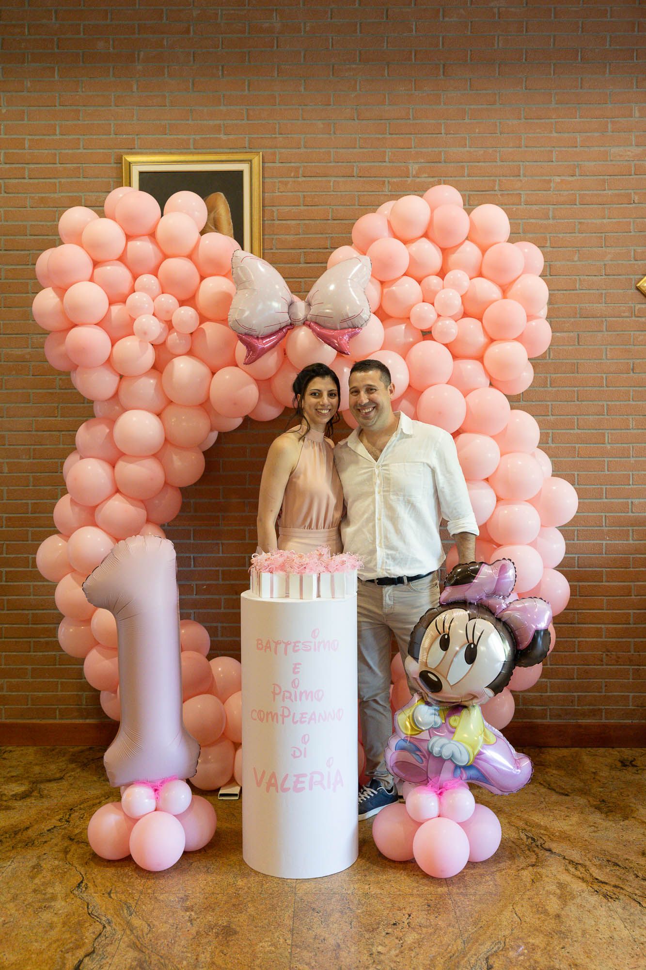 Couple posing at a baby girl first birthday party with pink balloon arch, Minnie Mouse balloon, and decorated cake.
